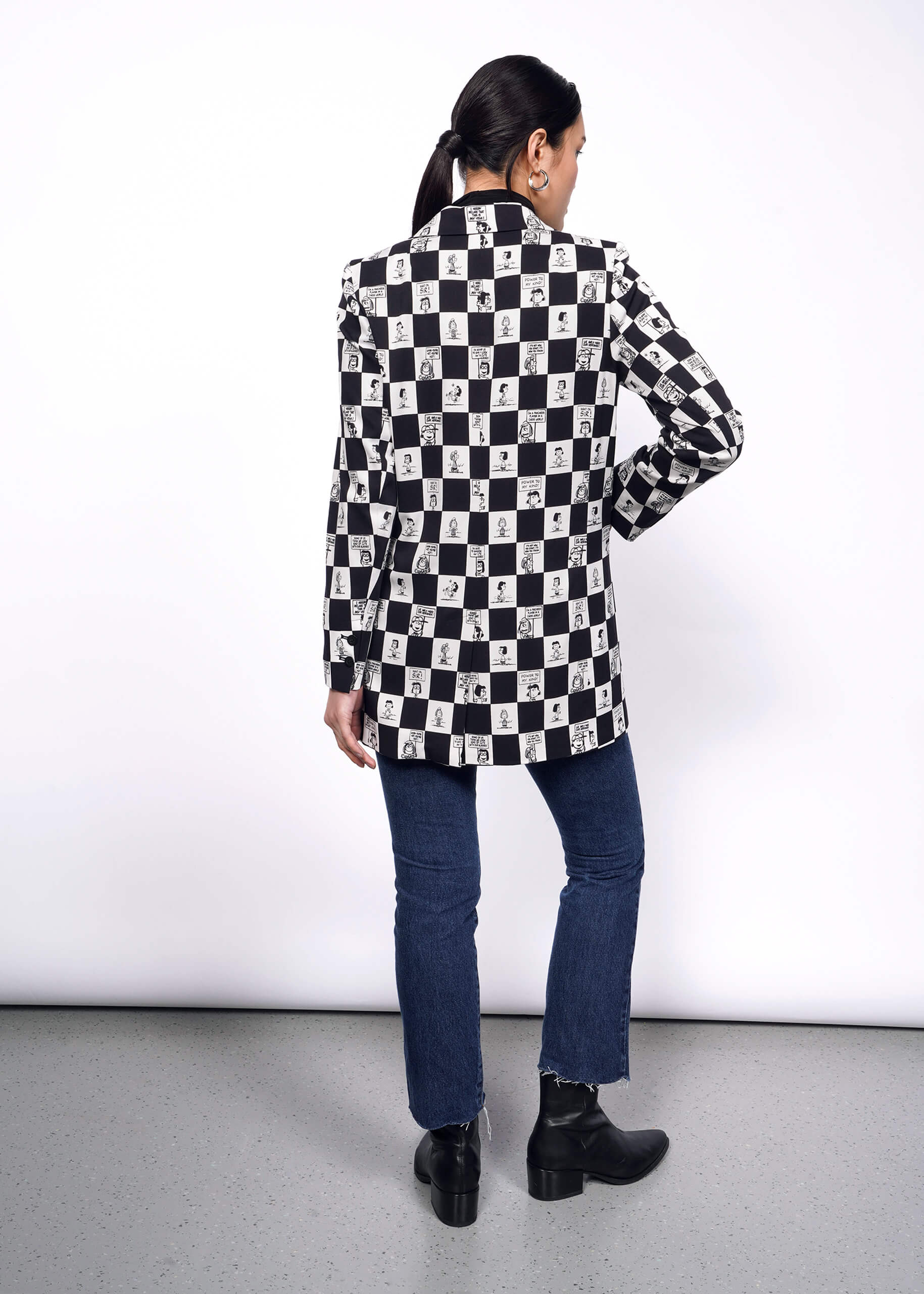 S=Back full body view of model with black hair tied back wearing jeans, and The Peanuts x Wildfang Double Breasted Blazer in a custom black and white checkered print featuring Peanuts characters.