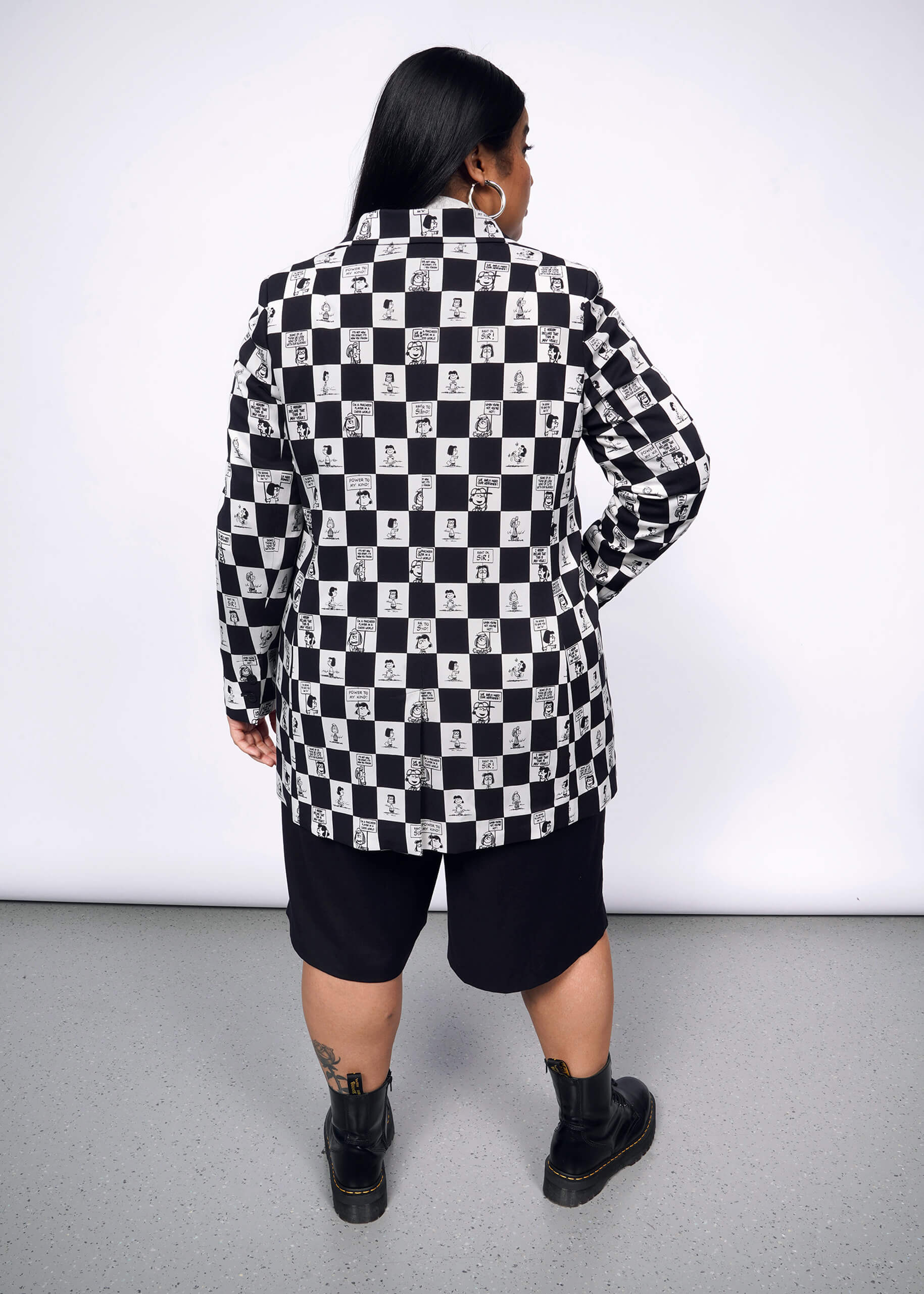 1X=Back view of person with long black hair wearing The Empower Wide Leg Short in black, and The Peanuts x Wildfang Double Breasted Blazer in a custom black and white checkered print featuring Peanuts characters, with black boots.