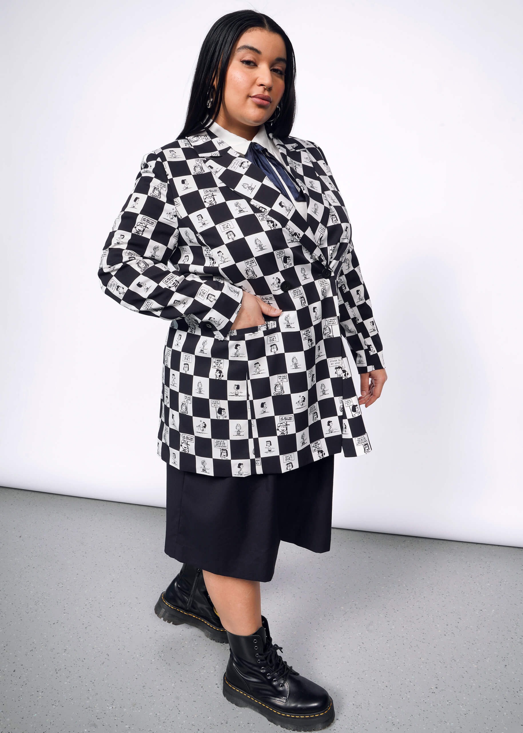 1X=Side view of person with long black hair wearing The Empower Button Up, The Empower Wide Leg Short, and The Peanuts x Wildfang Double Breasted Blazer in a custom black and white checkered print featuring Peanuts characters, with black boots.