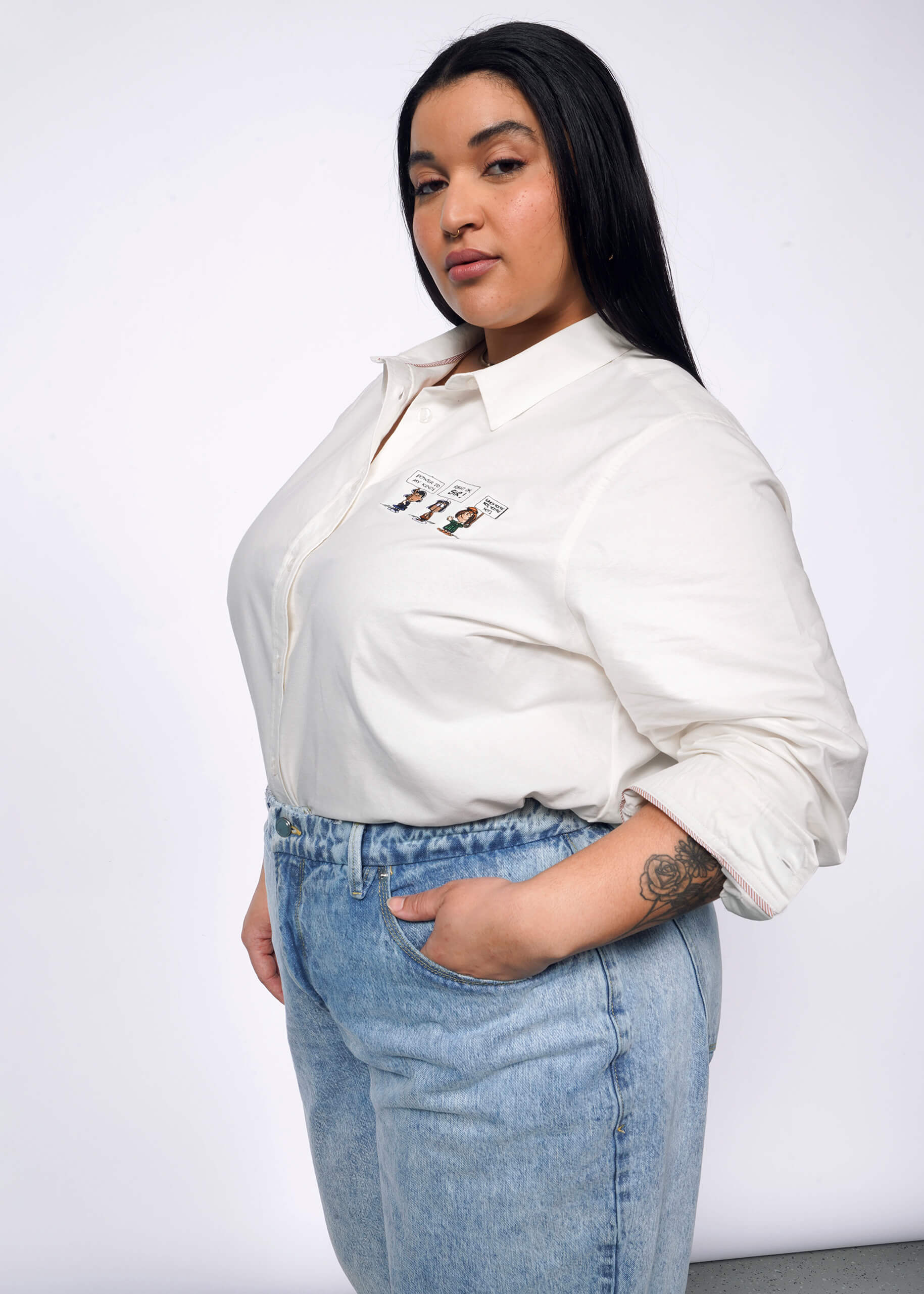 1X= Side view of person with long black hair looking at camera with hand in jeans pocket wearing The Peanuts x Wildfang Long Sleeve Button Up, which is a white shirt with Peanuts characters embroidered on left chest area.