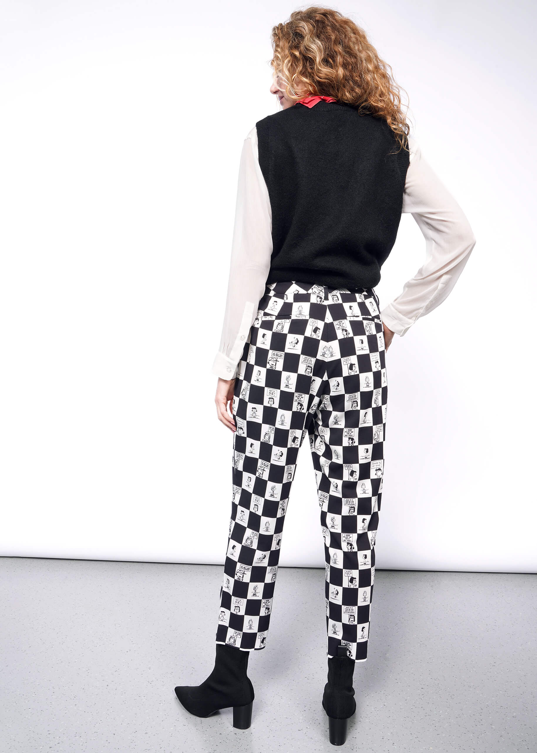 S=Back view of person with wavy blonde hair wearing white long sleeve button up under black sweater vest, a red bandana tied around their neck, black heeled boots, and the checkered black and white Slim Crop Pant from the Peanuts x Wildfang collection.