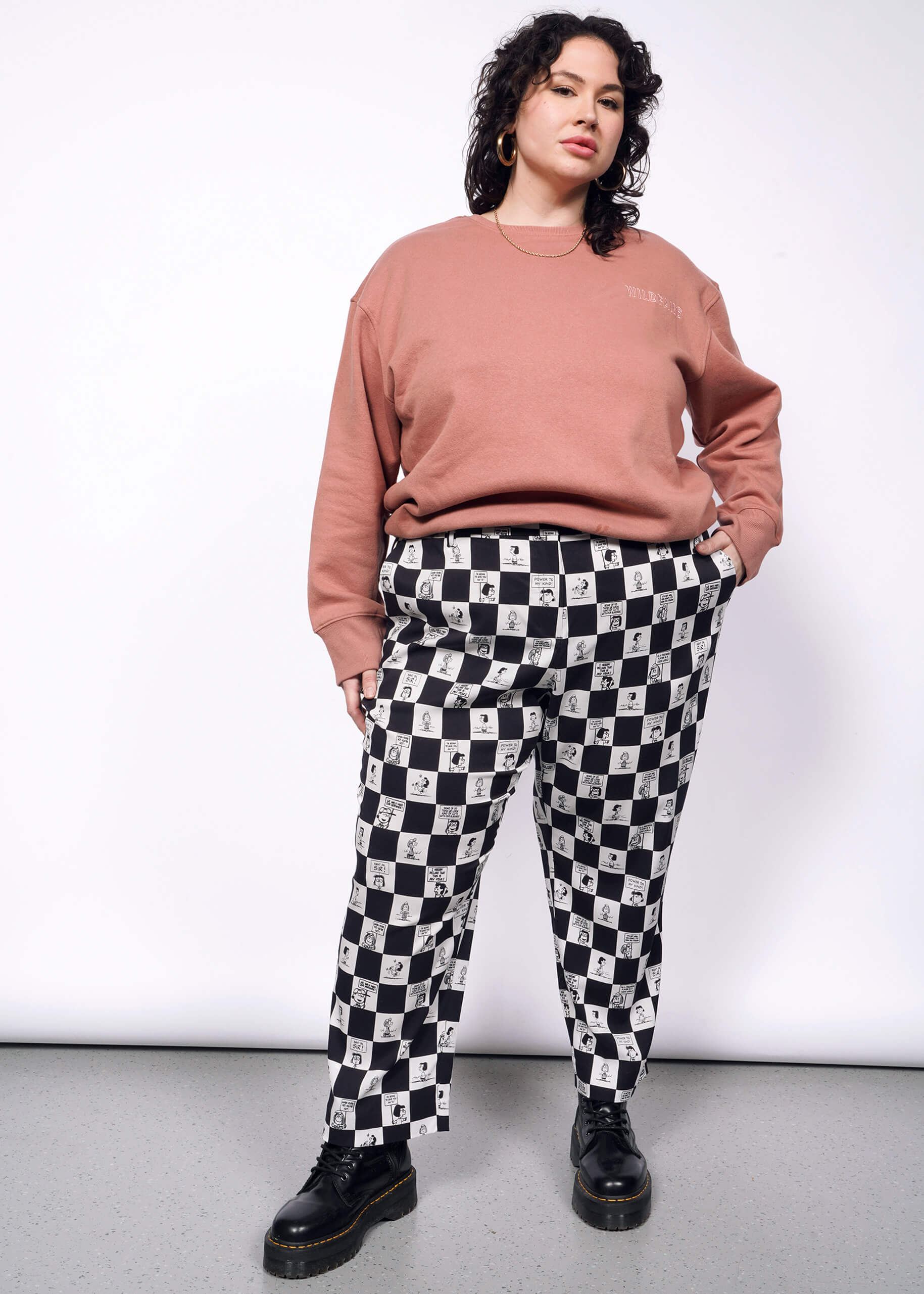 1X= Person with short curly black hair looks at camera wearing pink sweatshirt, black boots, and the checkered black and white Slim Crop Pant from the Peanuts x Wildfang collection.