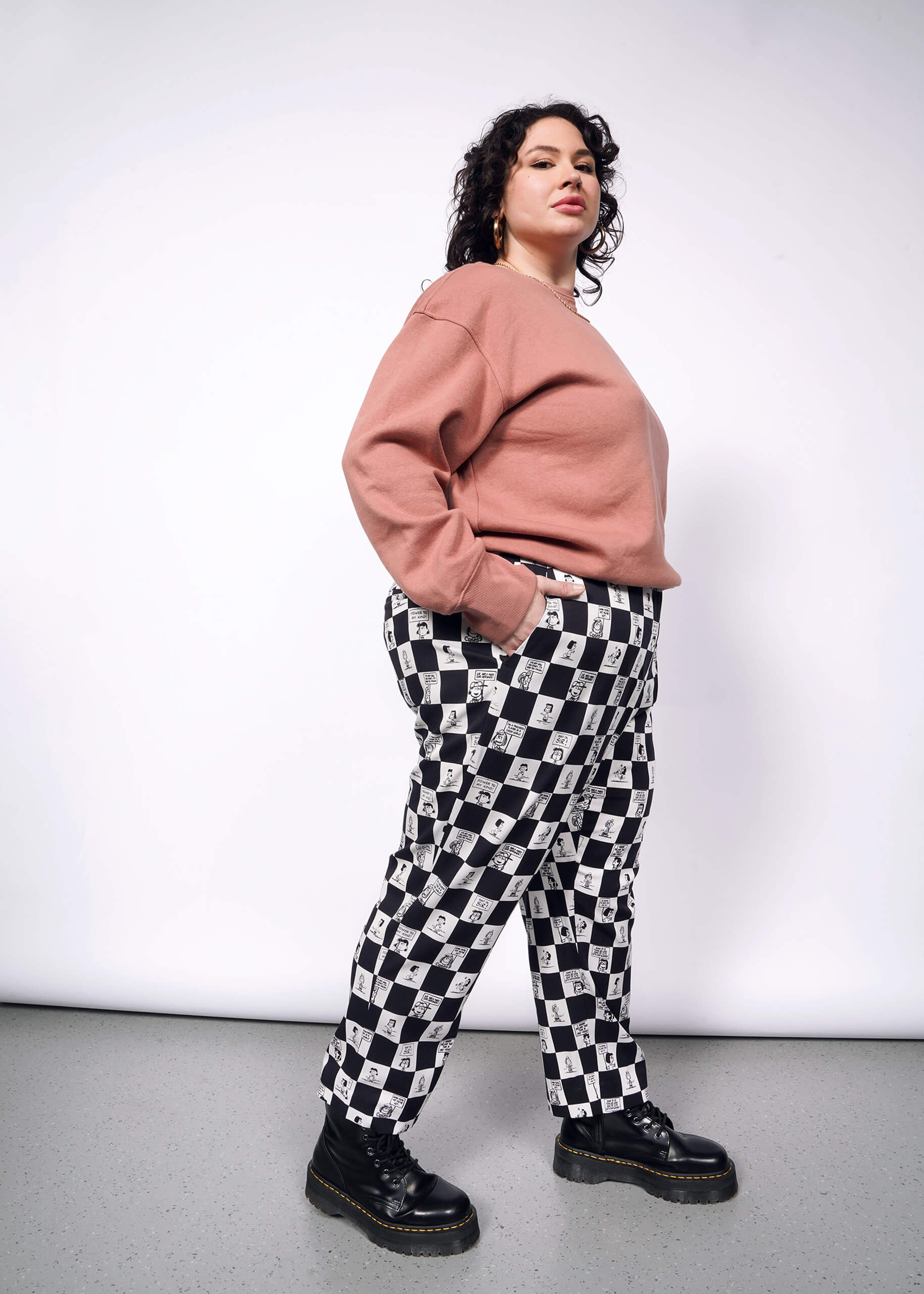 1X=Side view of person with short curly black hair looking at camera with hand in pocket while wearing pink sweatshirt, black boots, and the checkered black and white Slim Crop Pant from the Peanuts x Wildfang collection.