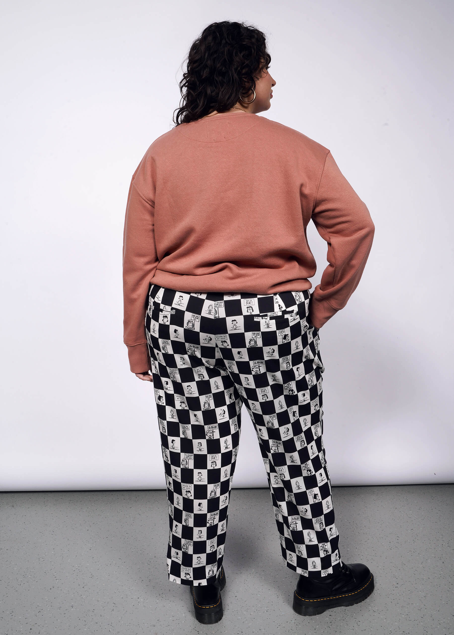 1X=Back view of person with short curly black hair looking at camera with hand in pocket while wearing pink sweatshirt, black boots, and the checkered black and white Slim Crop Pant from the Peanuts x Wildfang collection.