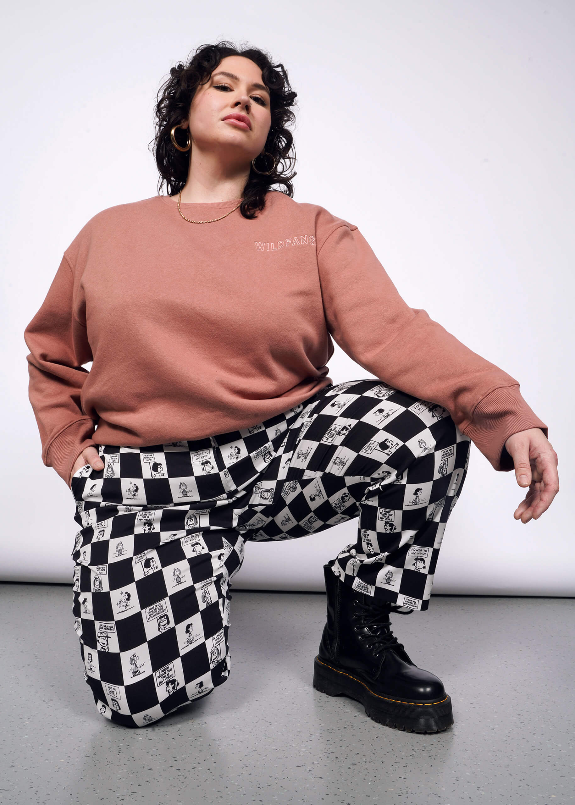 1X= Person with short curly black hair bends down on one knee wearing pink sweatshirt, black boots, and the checkered black and white Slim Crop Pant from the Peanuts x Wildfang collection.