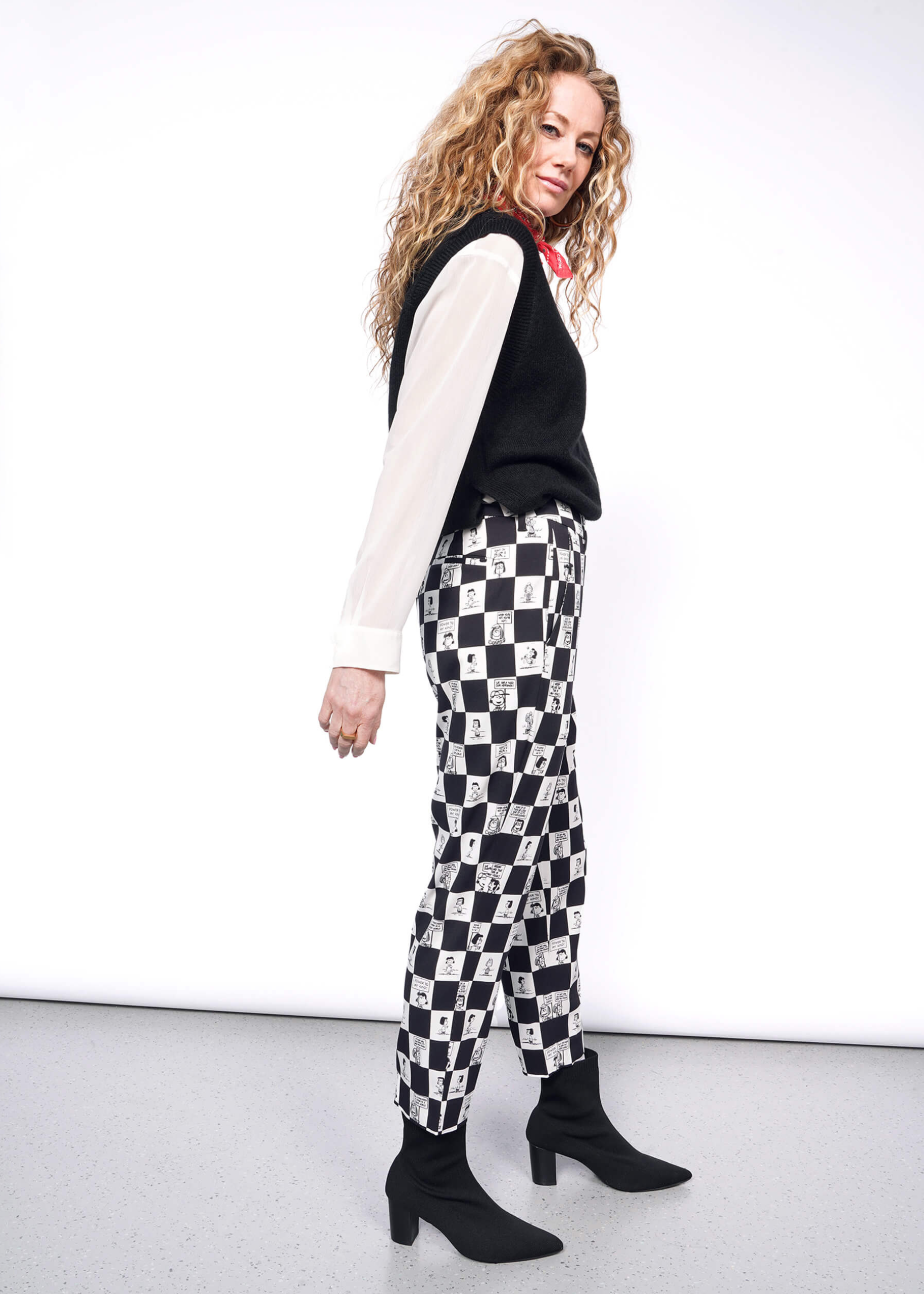 S=Side view of person with wavy blonde hair looking at camera wearing white long sleeve button up under black sweater vest, a red bandana tied around their neck, black heeled boots, and the checkered black and white Slim Crop Pant from the Peanuts x Wildfang collection.