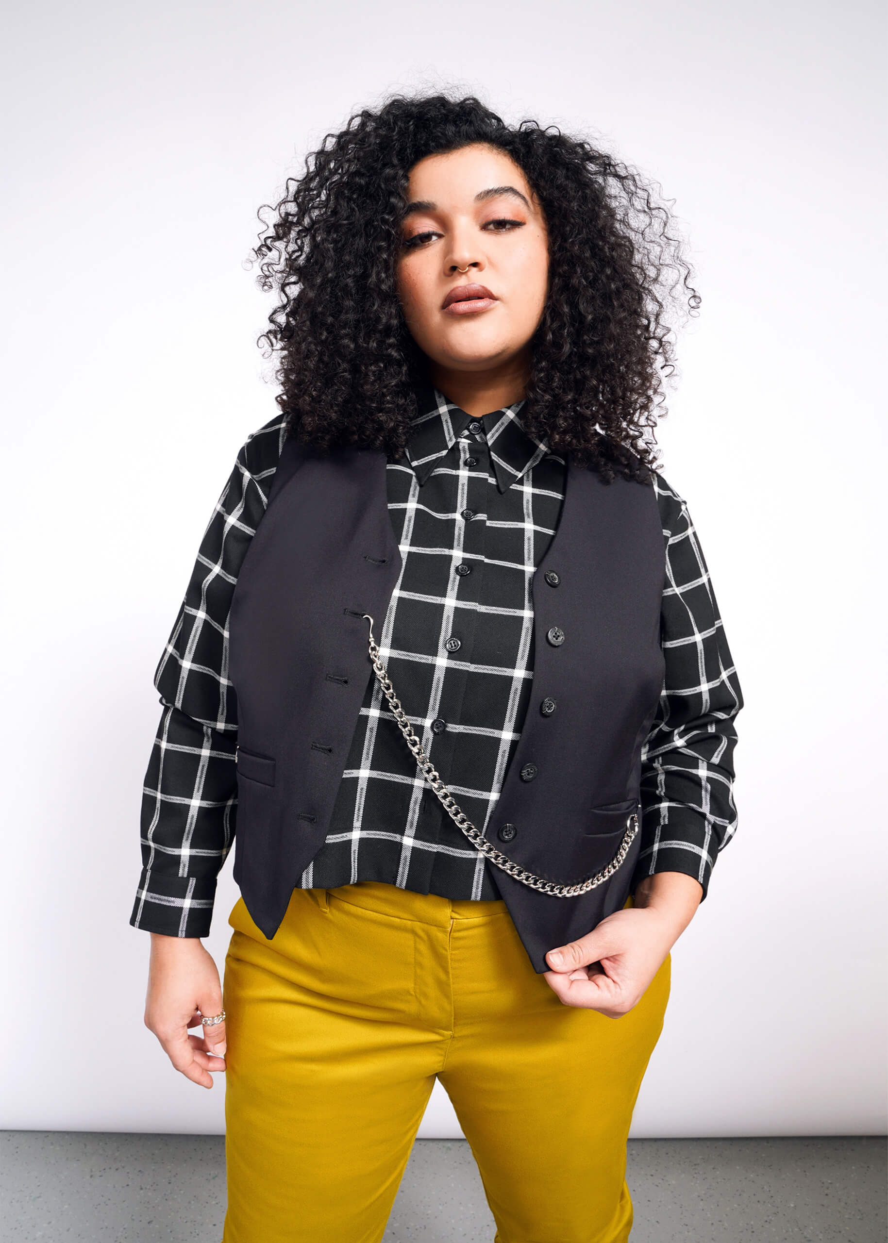 1X=A curly-haired person models a Debbie Harry X Wildfang Vest over a black and white checkered shirt with mustard yellow trousers, set against a plain backdrop.
