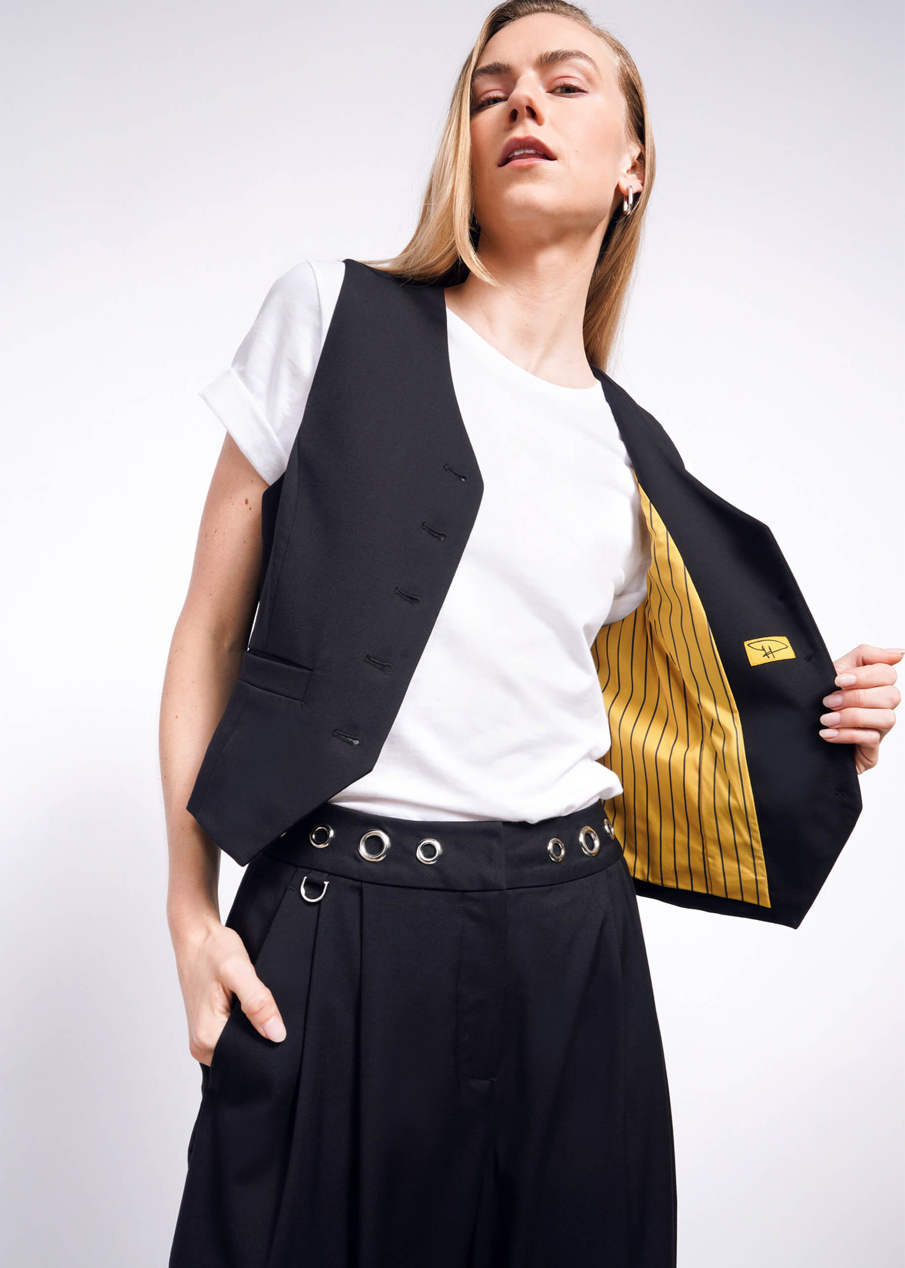 S=A person confidently poses in a white t-shirt, an open Debbie Harry X Wildfang Vest with yellow striped lining, and black grommet-adorned trousers against a plain background.