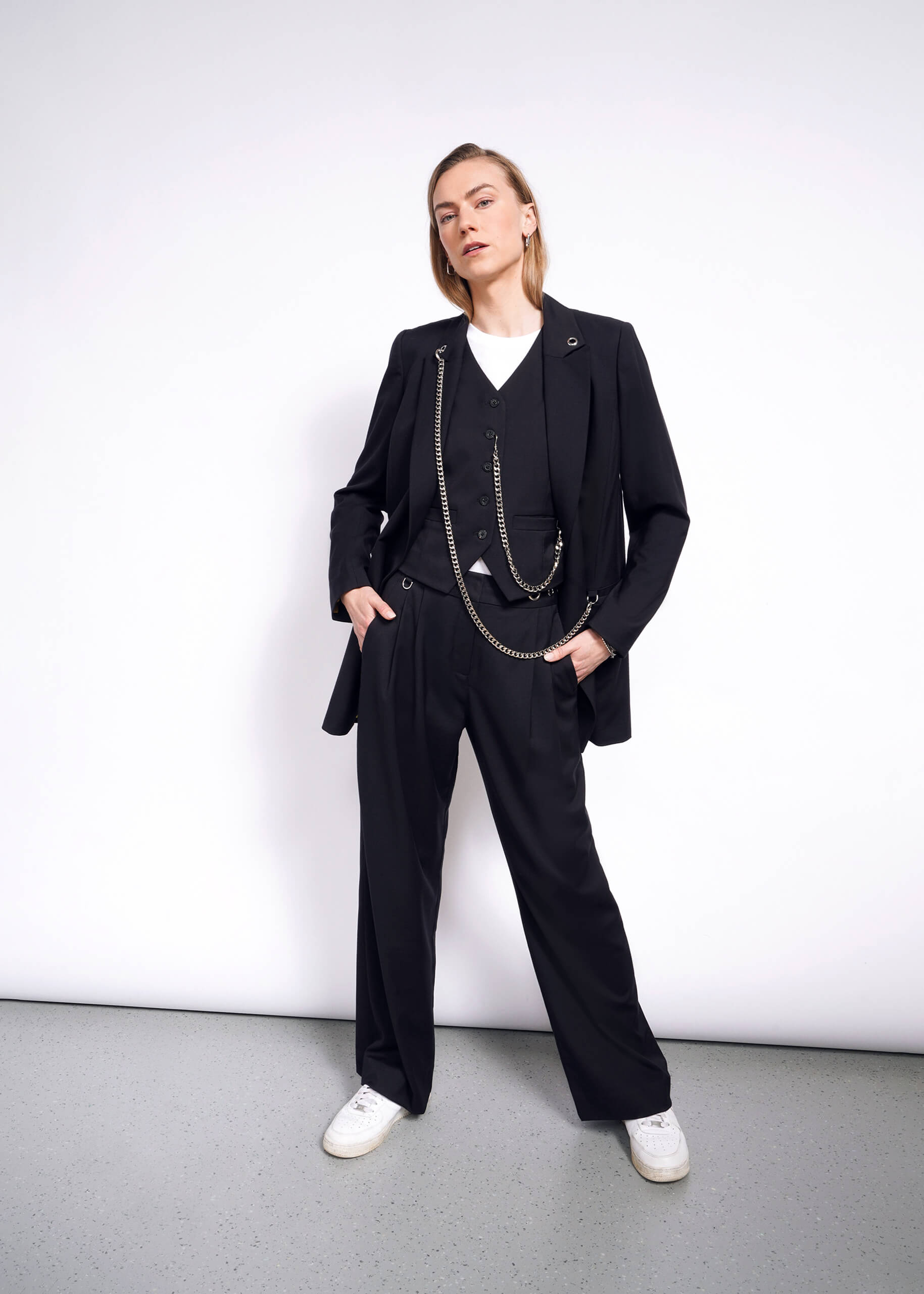 S=Posing against a white wall, the person wears a black suit with a Debbie Harry X Wildfang vest, white shirt, and sneakers. Hands in pockets, they strike a confident pose with a slight head tilt on the neutral gray floor.