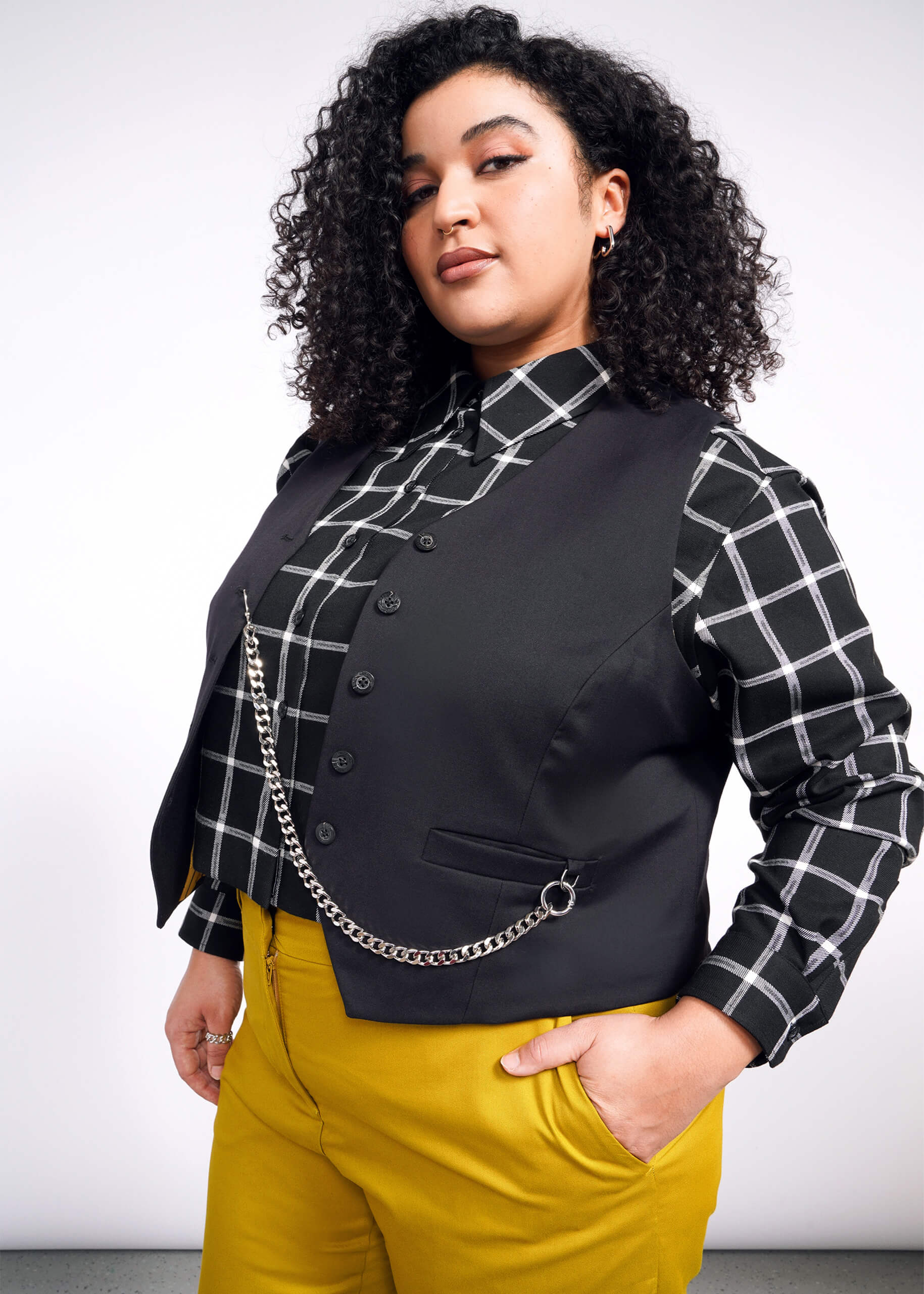1X=A person with curly hair, a black and white checkered shirt, a Debbie Harry X Wildfang Vest, and mustard yellow pants stands confidently against a simple background.