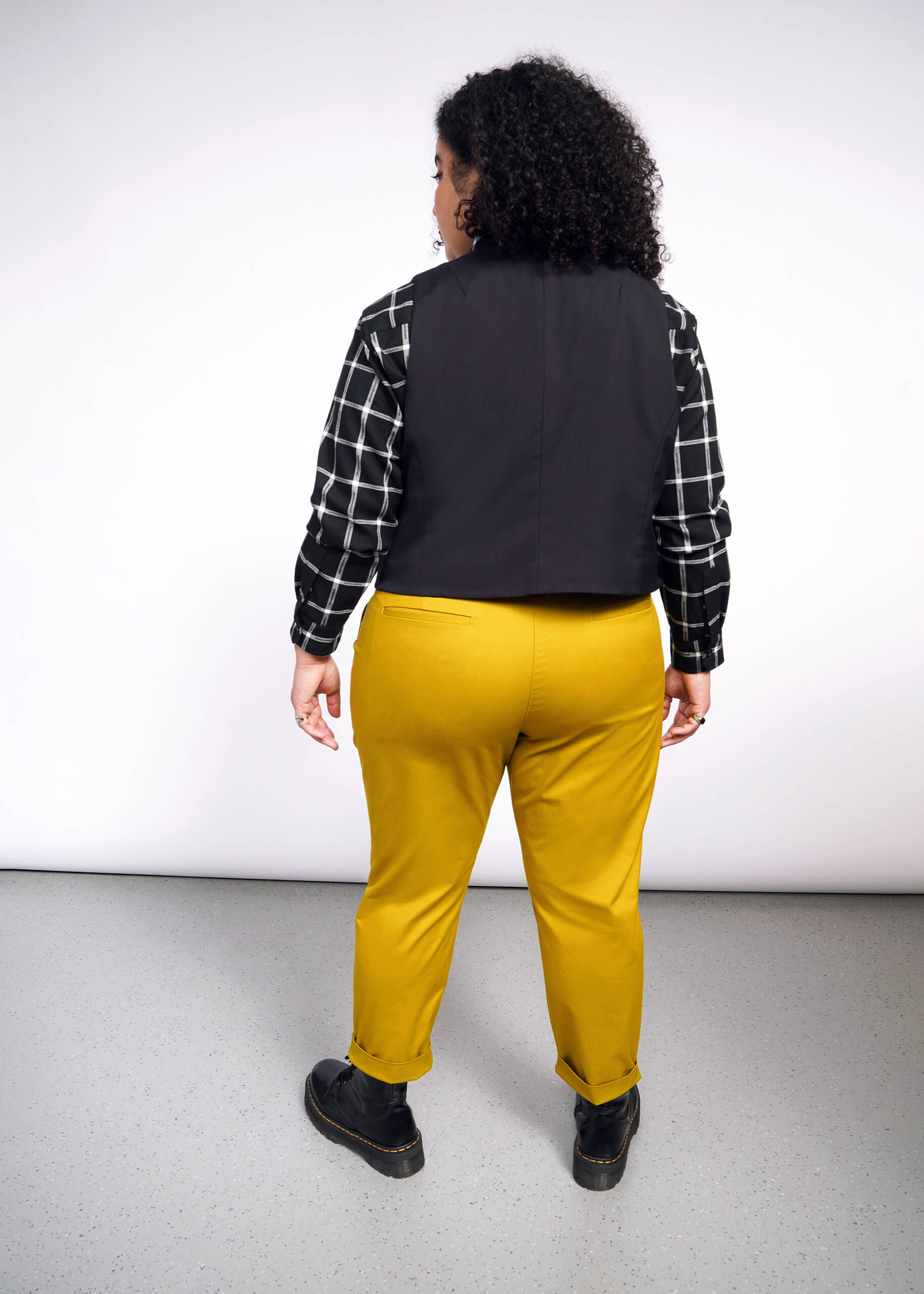 1X=Rear view of a person with curly hair in a checkered shirt, Debbie Harry X Wildfang Vest, mustard yellow pants, and black shoes against a plain light background.