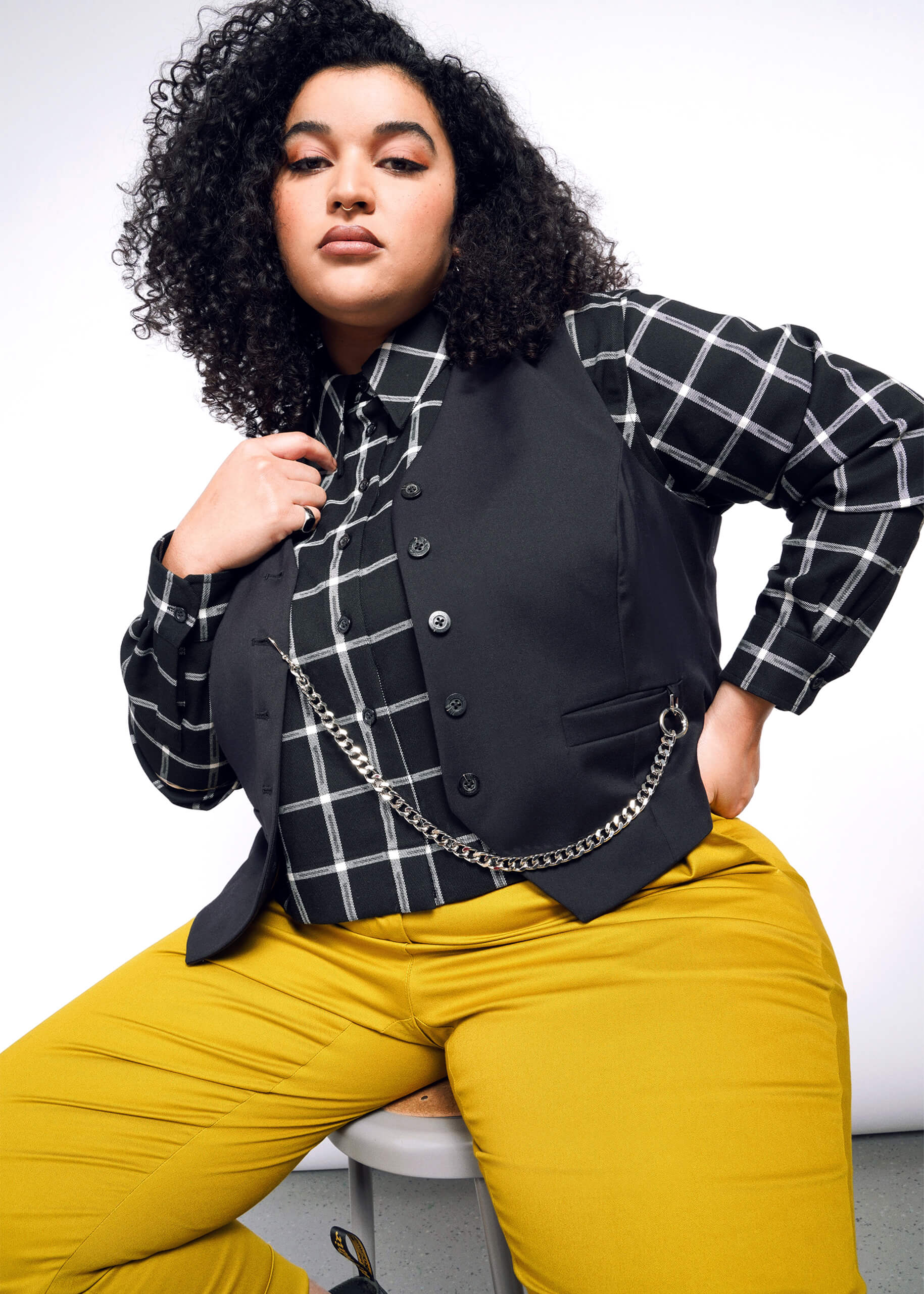 1X= The person with curly hair sits confidently on a stool against a white backdrop, wearing a black and white checkered shirt, bright yellow pants, and the Debbie Harry X Wildfang Vest enhanced with a chain.
