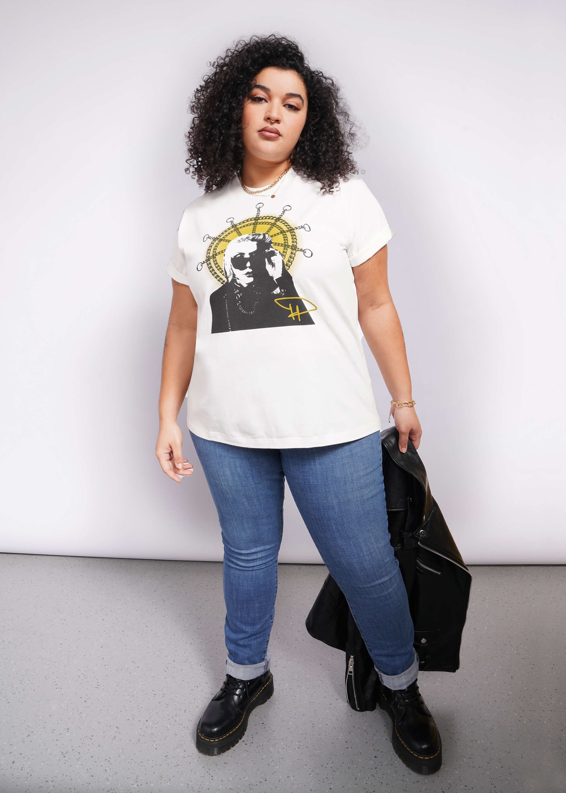1X=A person with curly hair confidently rocks a Debbie Harry X Wildfang Graphic Tee. Paired with blue jeans and black boots, they hold a black jacket against a plain backdrop, exuding an effortlessly cool vibe reminiscent of classic punk fashion.