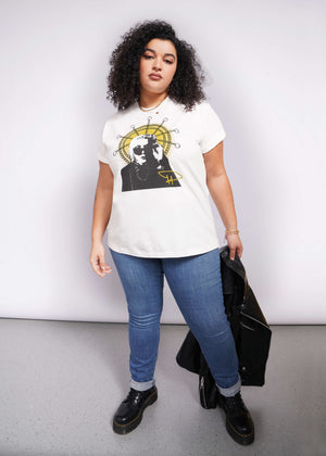 1X=A person with curly hair confidently rocks a Debbie Harry X Wildfang Graphic Tee. Paired with blue jeans and black boots, they hold a black jacket against a plain backdrop, exuding an effortlessly cool vibe reminiscent of classic punk fashion.