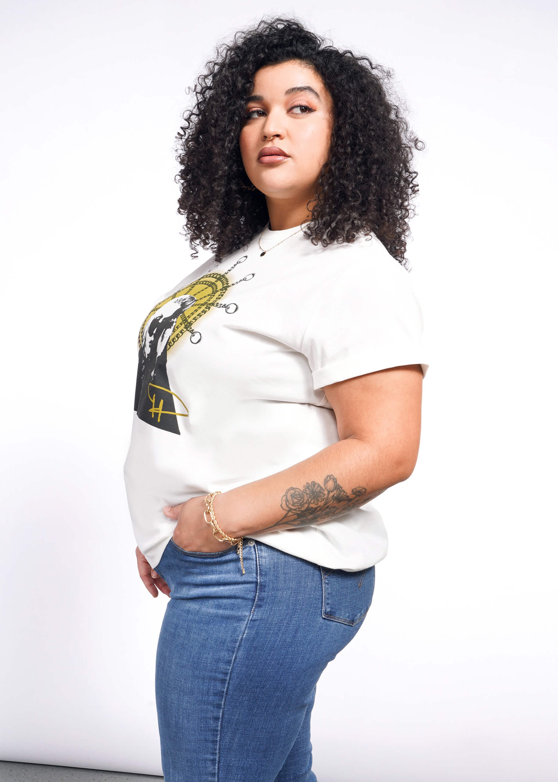 1X= A person with curly hair stands sideways, donning a Debbie Harry X Wildfang Graphic Tee and blue jeans. Their hand is in their pocket, showcasing an arm tattoo. A plain white background accentuates their punk fashion style.