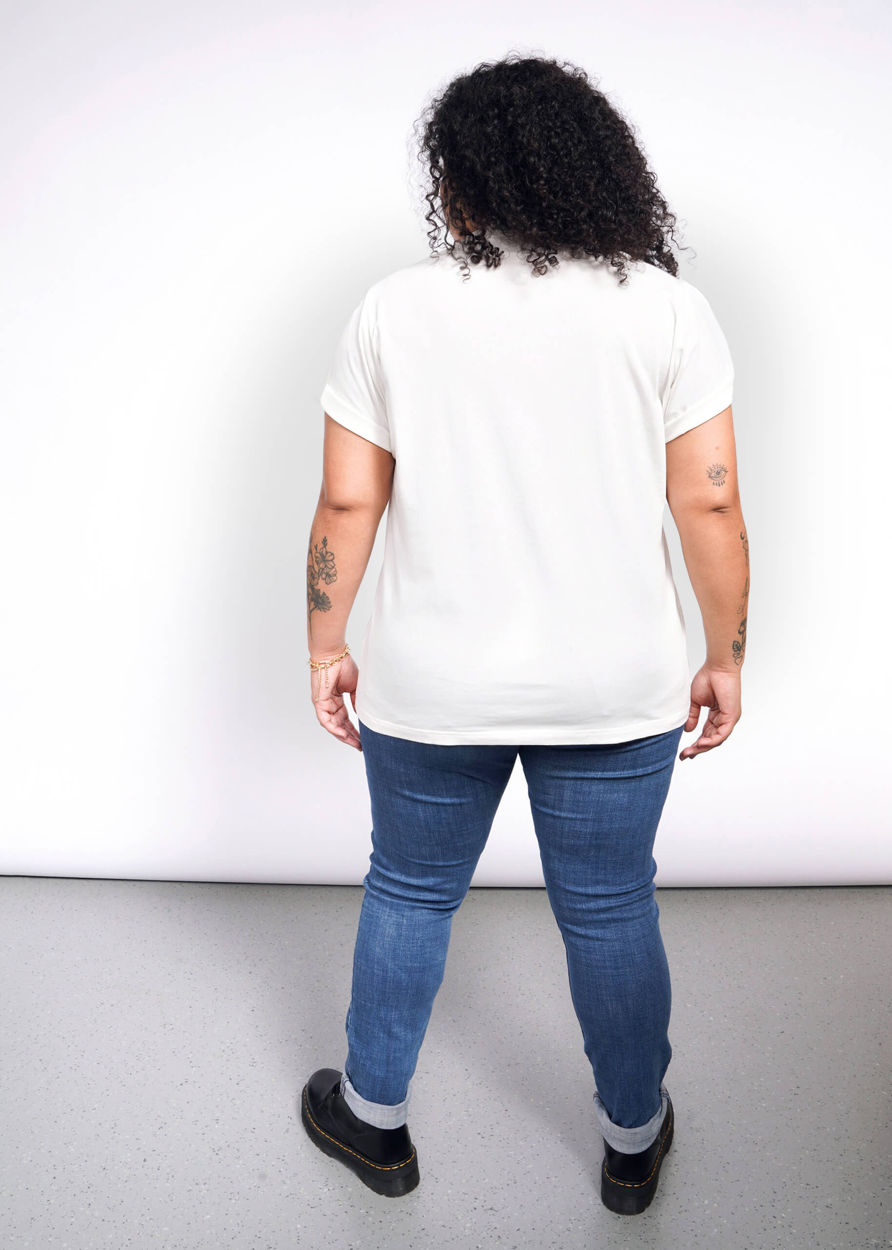 1X=A curly-haired person, tattoos visible, stands back turned on a speckled gray floor. Theyre in a Debbie Harry X Wildfang Graphic Tee, blue jeans, and black shoes against a plain white backdrop, exuding punk fashion vibes.