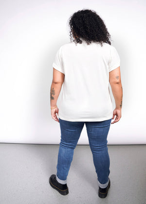 1X=A curly-haired person, tattoos visible, stands back turned on a speckled gray floor. Theyre in a Debbie Harry X Wildfang Graphic Tee, blue jeans, and black shoes against a plain white backdrop, exuding punk fashion vibes.
