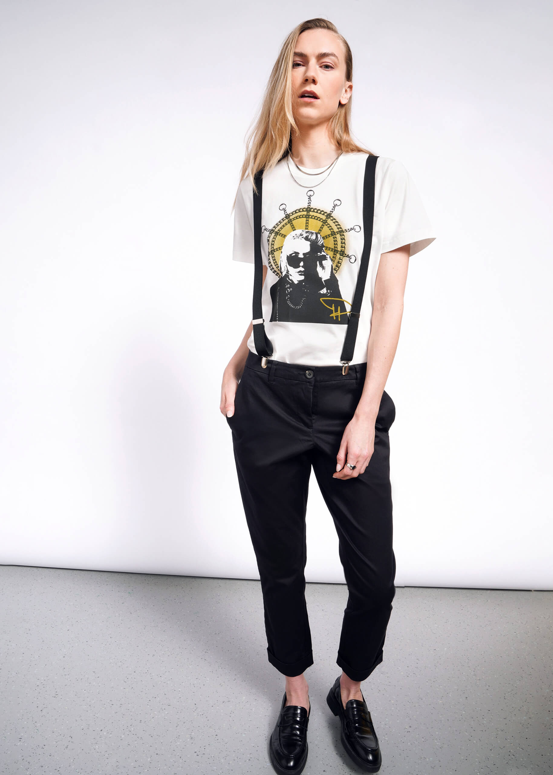 S=A person with long blond hair stands against a white background, wearing a Debbie Harry X Wildfang Graphic Tee. Black suspenders, pants, and loafers complete the punk look as they maintain a neutral expression with hands in their pockets.