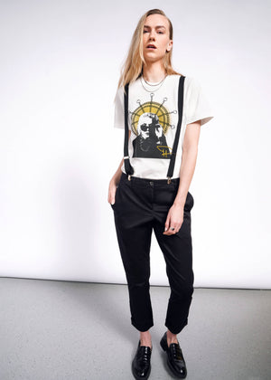 S=A person with long blond hair stands against a white background, wearing a Debbie Harry X Wildfang Graphic Tee. Black suspenders, pants, and loafers complete the punk look as they maintain a neutral expression with hands in their pockets.
