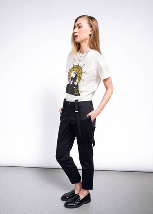 S= A person with long hair dons the Debbie Harry X Wildfang Graphic Tee, paired with black pants and suspenders. Against a light gray wall, hands in pockets, their shiny black shoes shine under the light, capturing punk fashion effortlessly.