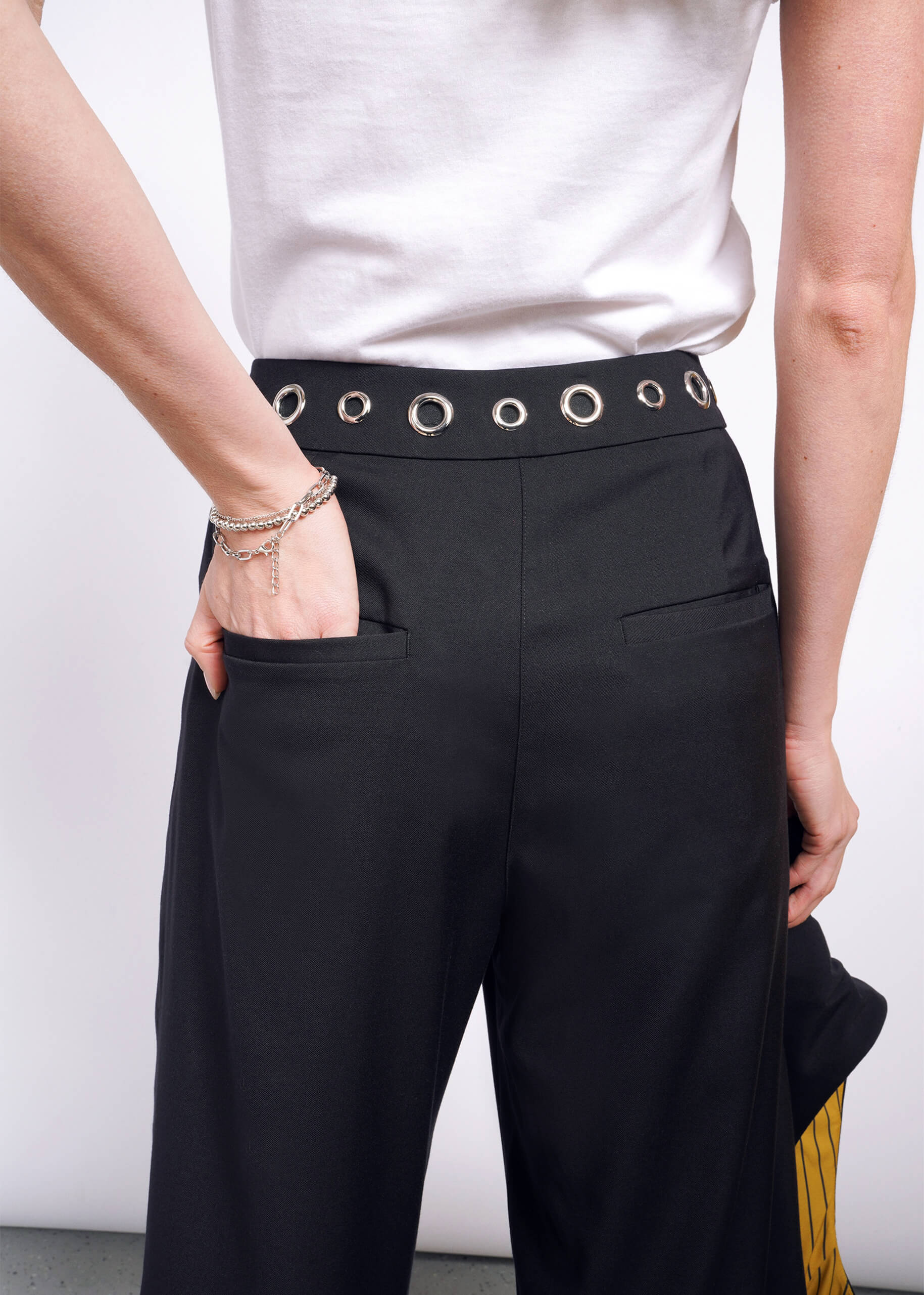 S=A person is wearing a white shirt and the Debbie Harry X Wildfang Wide Leg Trouser with silver eyelet waist details. Their left hand is in a pocket of the high-waisted pants, paired with a silver bracelet, standing out against a plain white background.