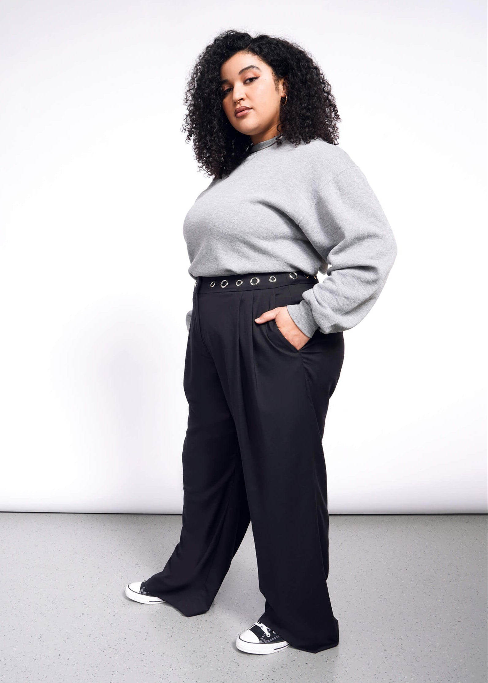 1X=Curly-haired and confident, a person poses against a white background in Debbie Harry X Wildfang Wide Leg Trousers, paired with a grey sweater and black sneakers. With hands in pockets, they look at the camera with a relaxed posture.