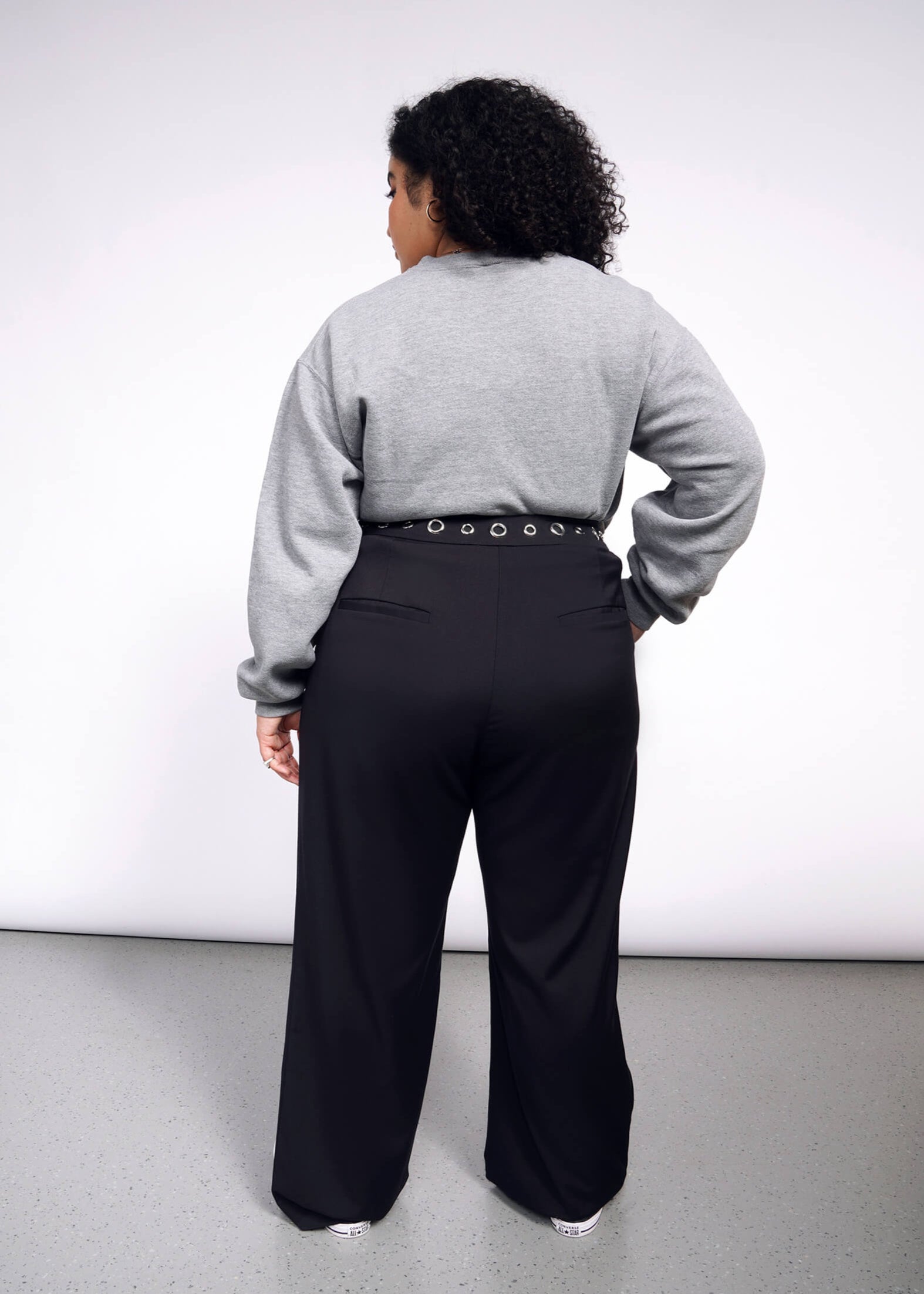1X= A person with curly hair faces away from the camera, wearing a gray sweater and black Debbie Harry X Wildfang Wide Leg Trouser. The minimalist backdrop features a gray floor, highlighting their chic ensemble.
