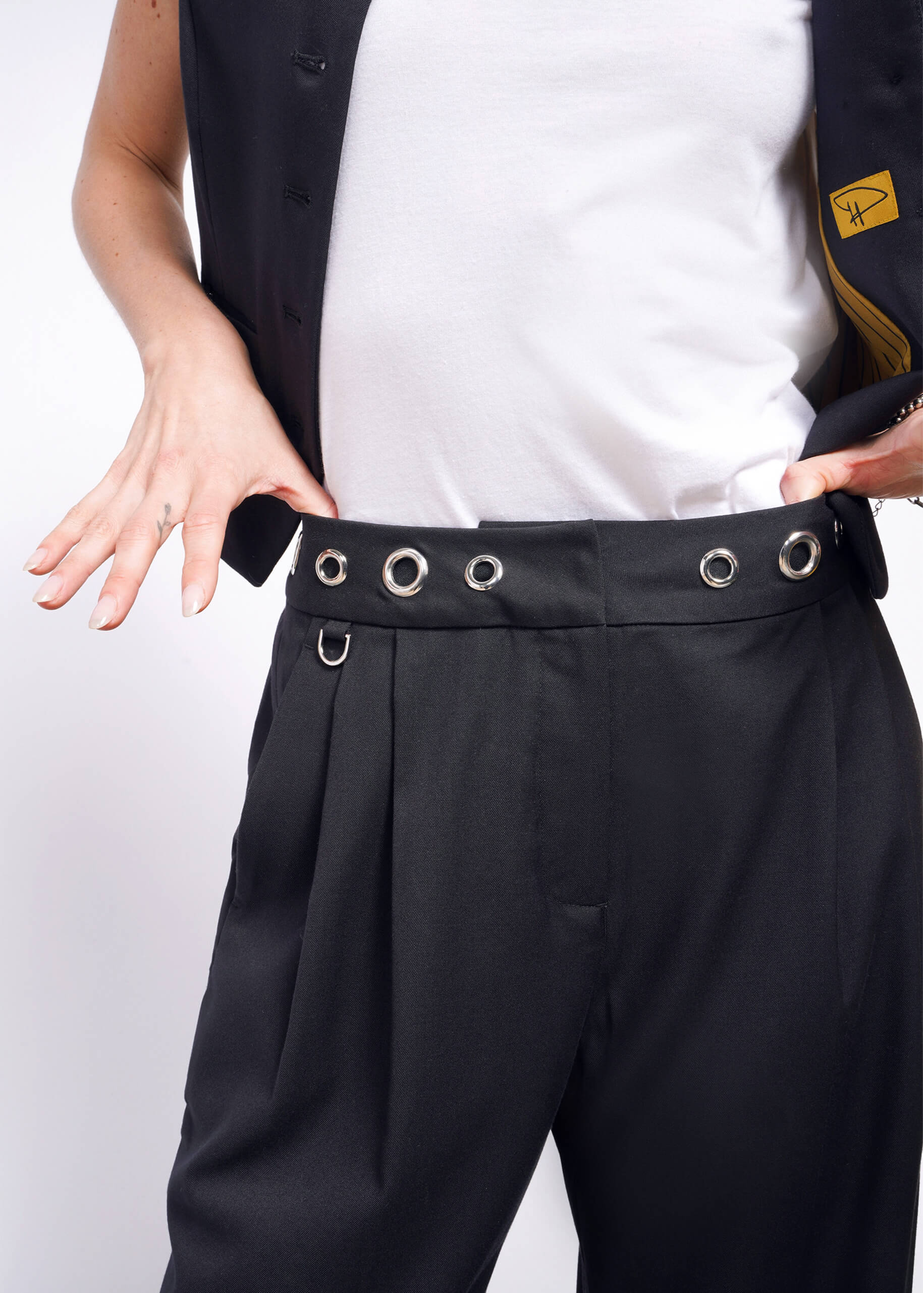 S= Theyre donning chic black Debbie Harry X Wildfang Wide Leg Trousers and a white shirt paired with a black sleeveless vest. The high-waisted fit with large metal eyelets enhances their silhouette, and their hands rest on the waist, revealing part of a left hand tattoo.