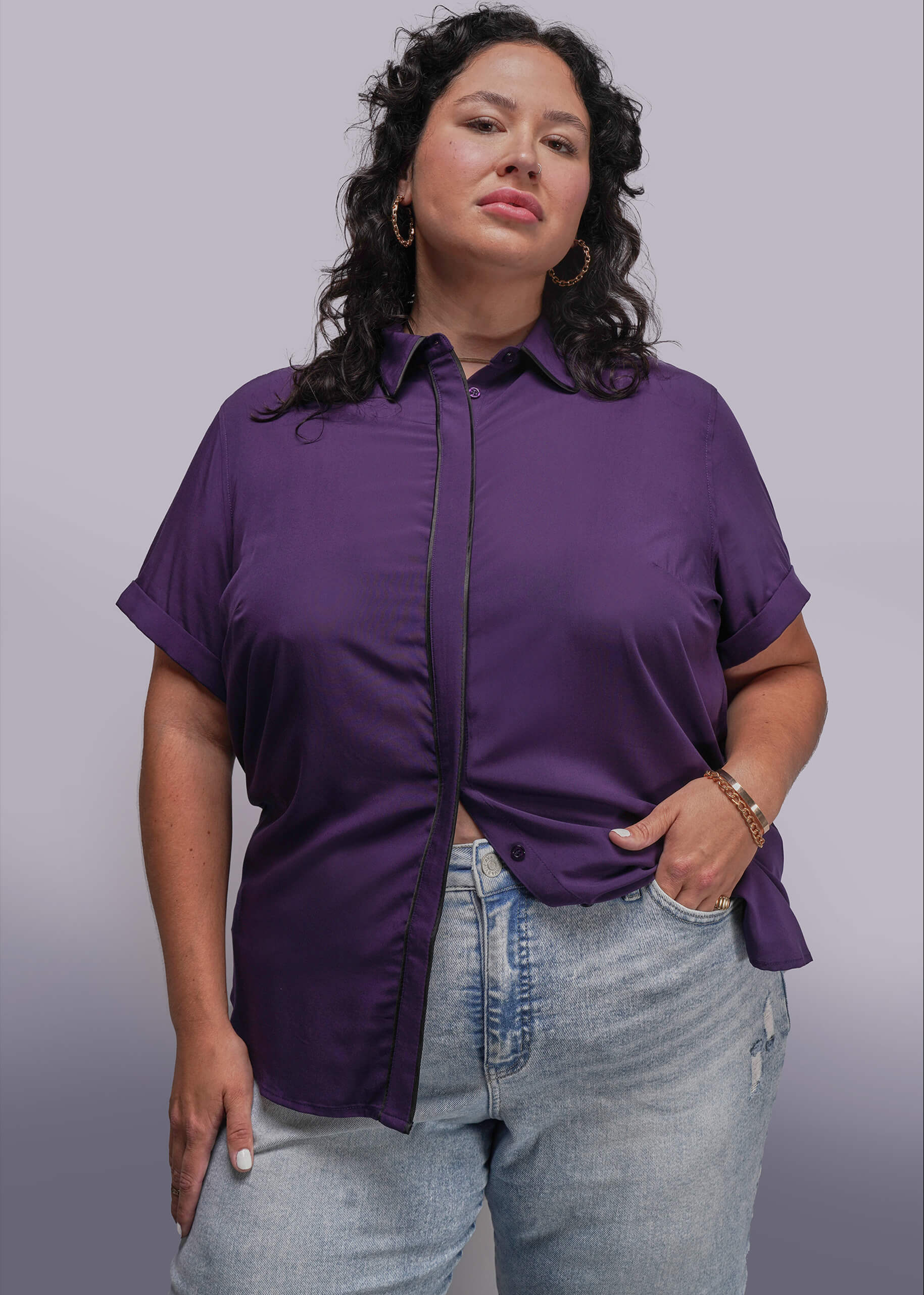 1X=Woman wearing a purple shirt and light blue jeans against a gray background