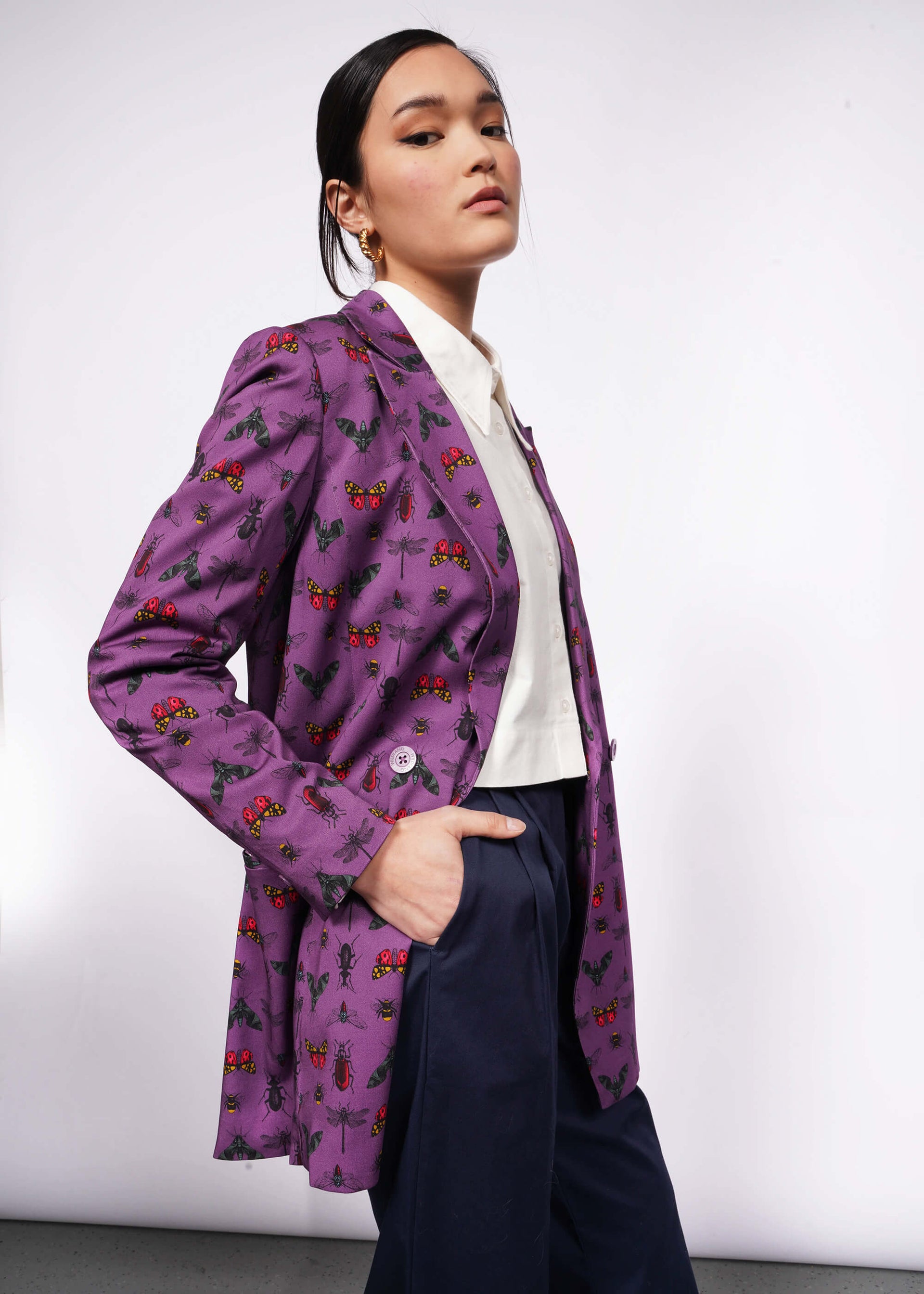 S=Side view of person with black hair and hand in trouser pocket wearing purple bug printed blazer unbuttoned with white button up shirt underneath