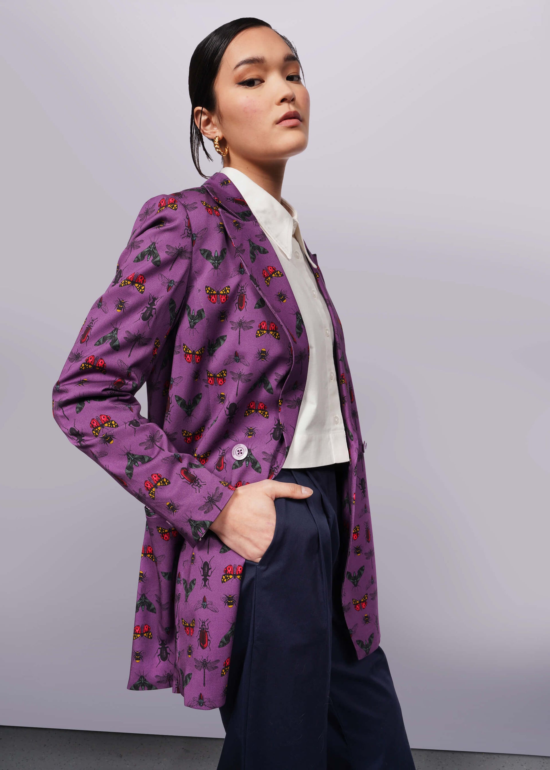 S=Side view of person with black hair and hand in trouser pocket wearing purple bug printed blazer unbuttoned with white button up shirt underneath