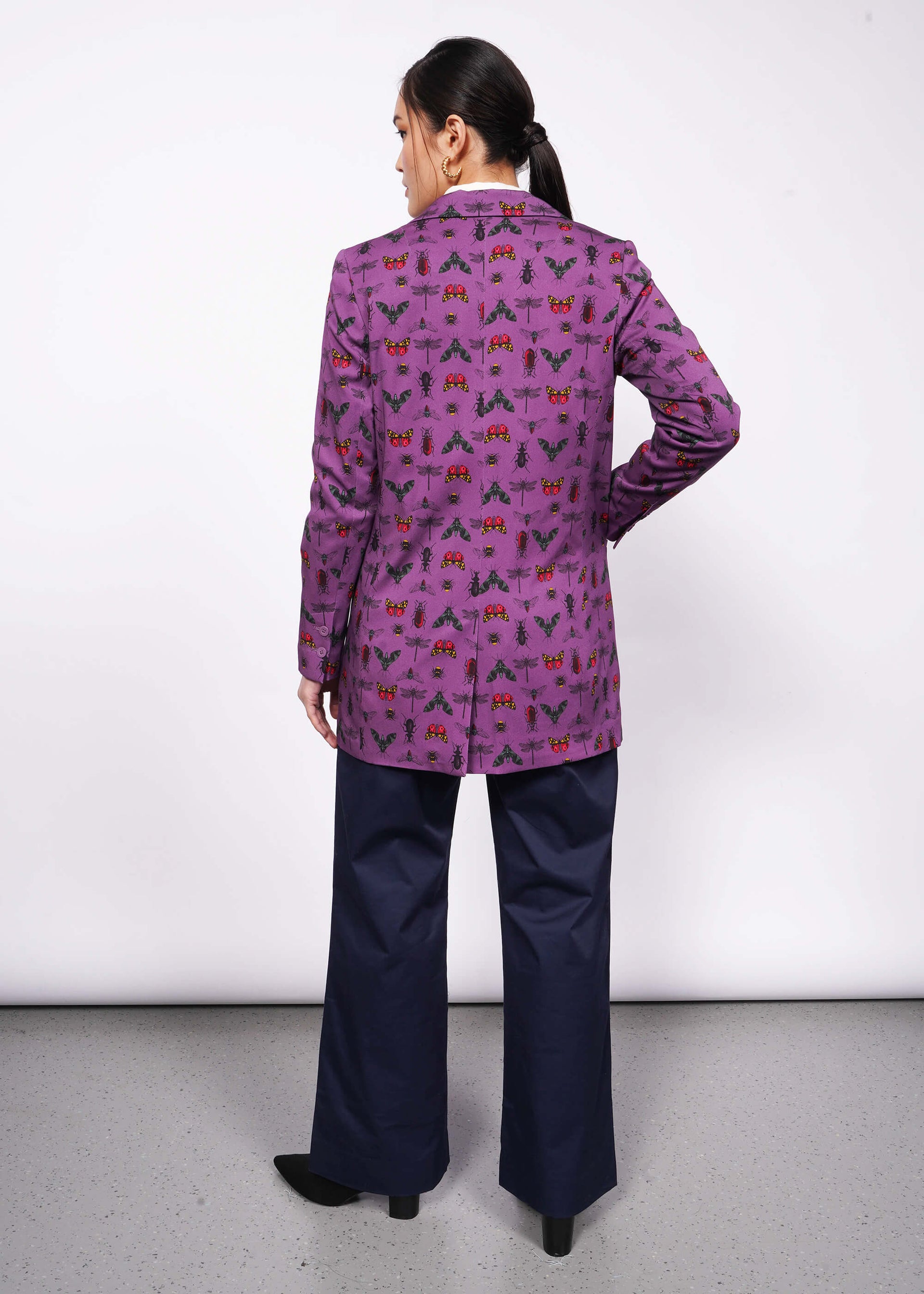 S=Back view of person with black hair and hand in trouser pocket wearing purple bug printed blazer with navy trouser and black heeled boots.