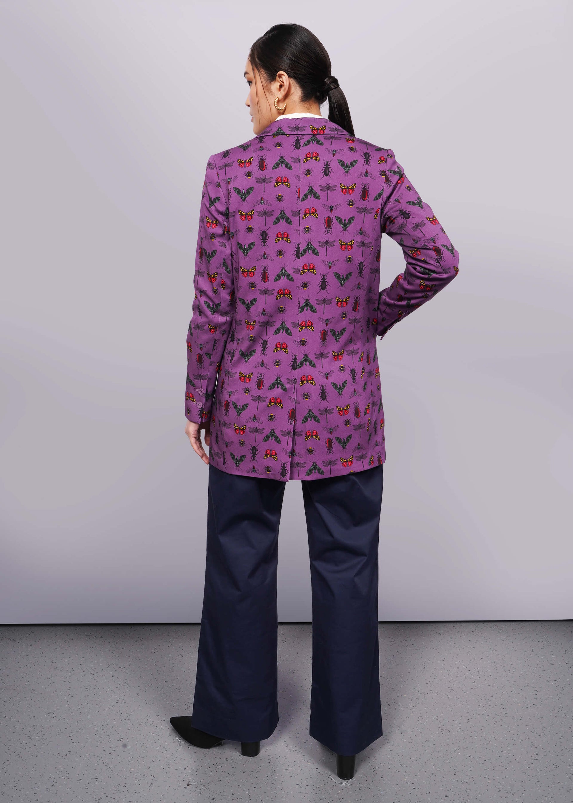 S=Back view of person with black hair and hand in trouser pocket wearing purple bug printed blazer with navy trouser and black heeled boots.