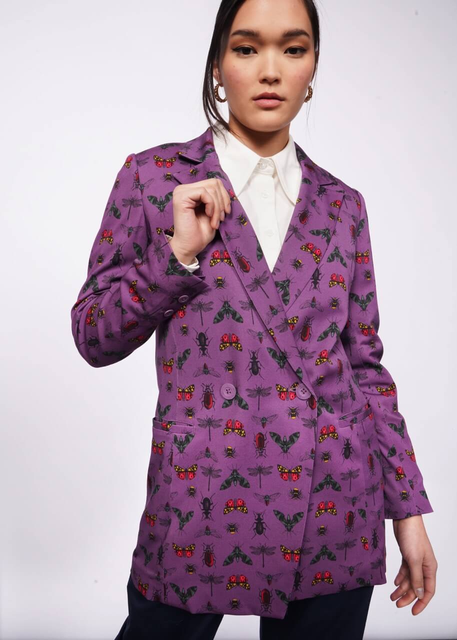 S= Person with black hair looks at camera holding the lapel of their purple blazer with a bug print on it. They are wearing a white shirt underneath. Neutral background.