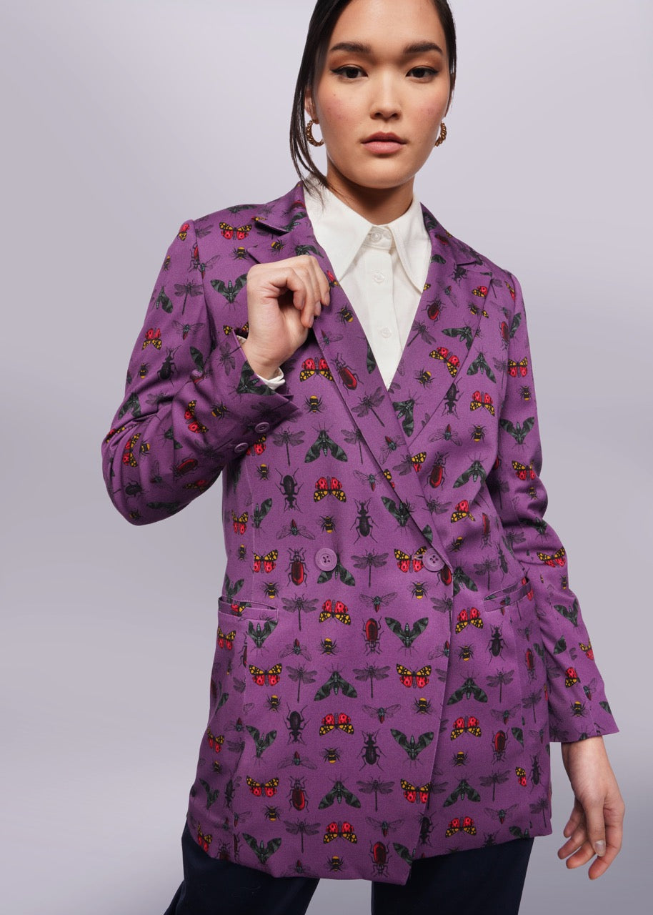 S= Person with black hair looks at camera holding the lapel of their purple blazer with a bug print on it. They are wearing a white shirt underneath. Neutral background.