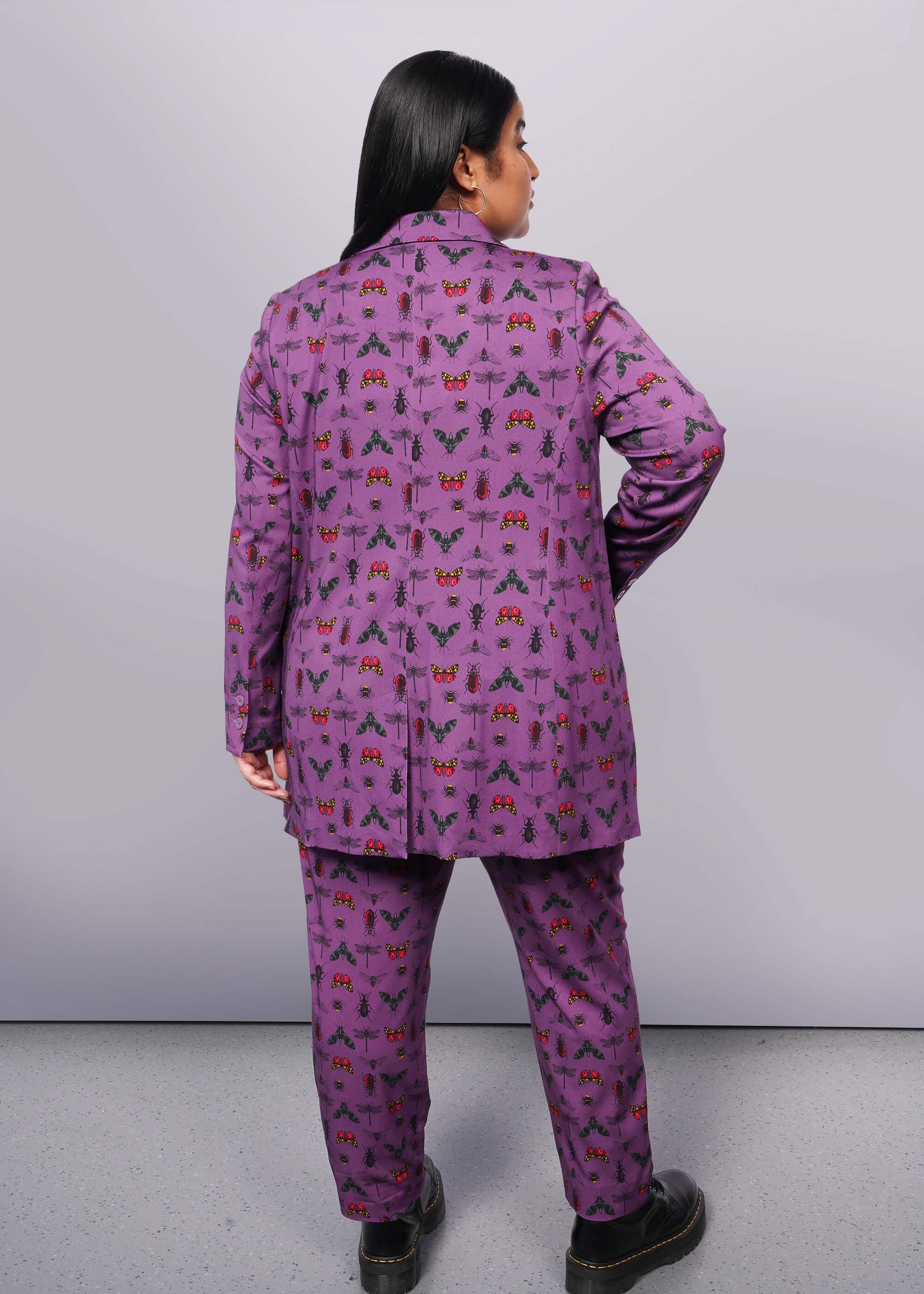 1X=Back view of person with long black hair with hand in double breasted blazer pocket. Is wearing matching trouser and black boots.