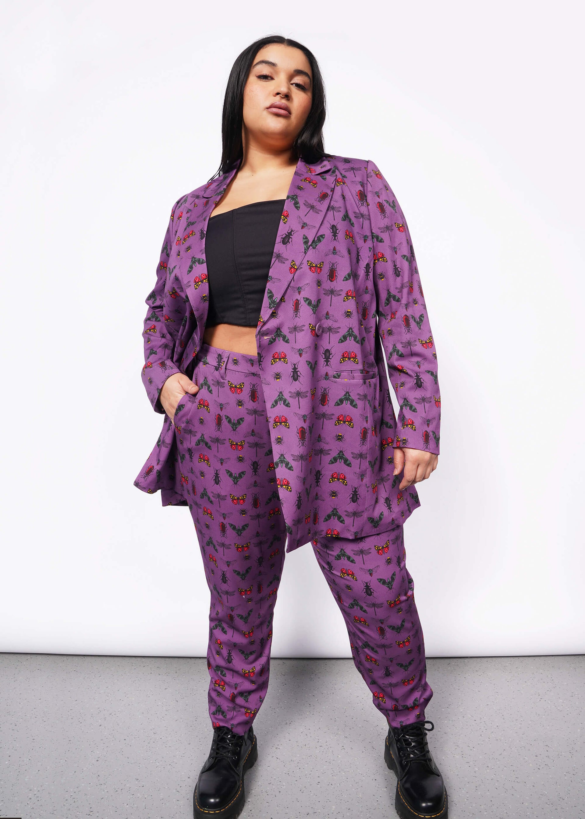 1X= Front view of person with long black hair and nose ring looking at camera with hand in pant pocket. Blazer is unbuttoned and is purple with bug print. They are wearing black corset underneath blazer, a matching pant, and a black boot.