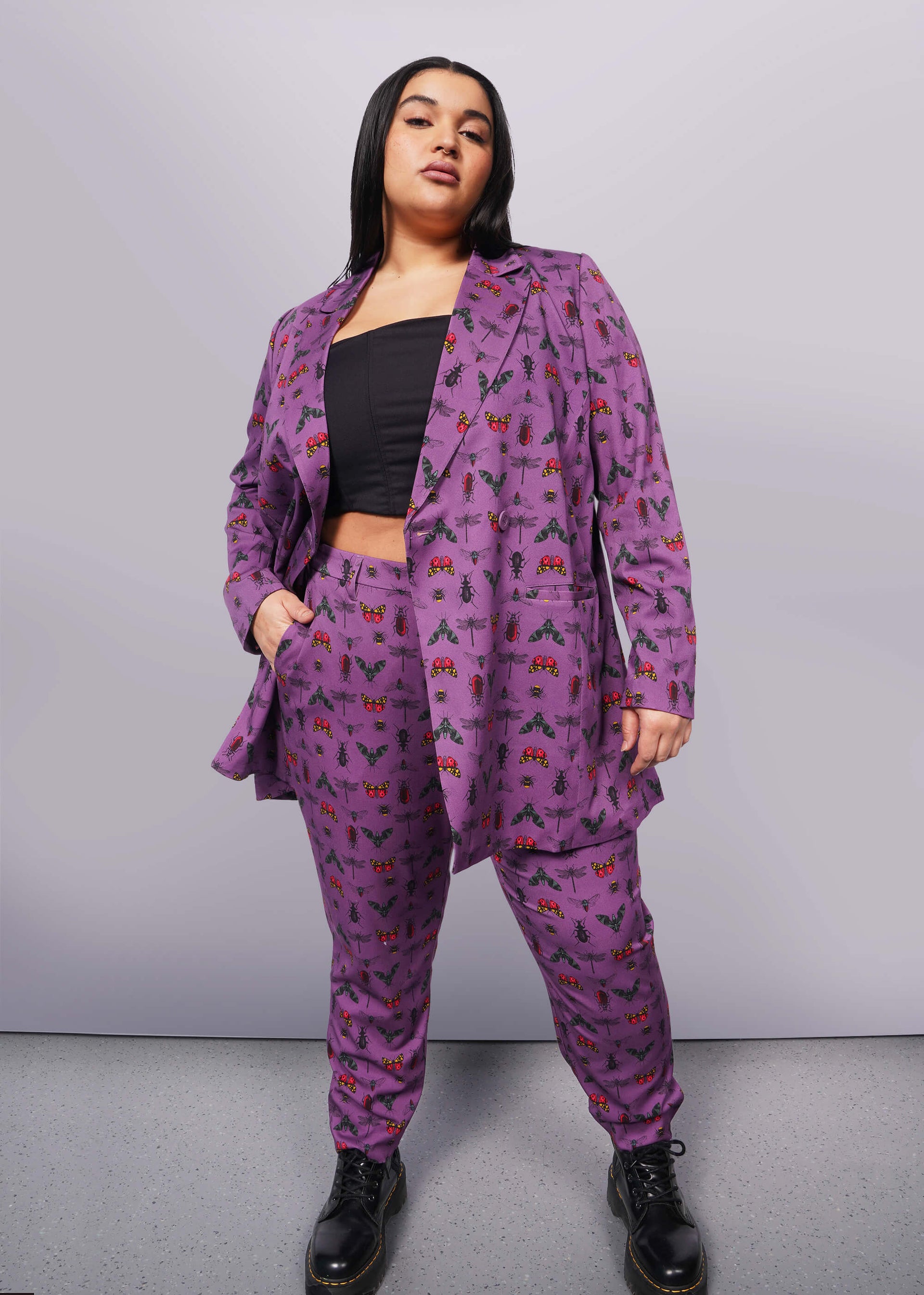 1X= Front view of person with long black hair and nose ring looking at camera with hand in pant pocket. Blazer is unbuttoned and is purple with bug print. They are wearing black corset underneath blazer, a matching pant, and a black boot.