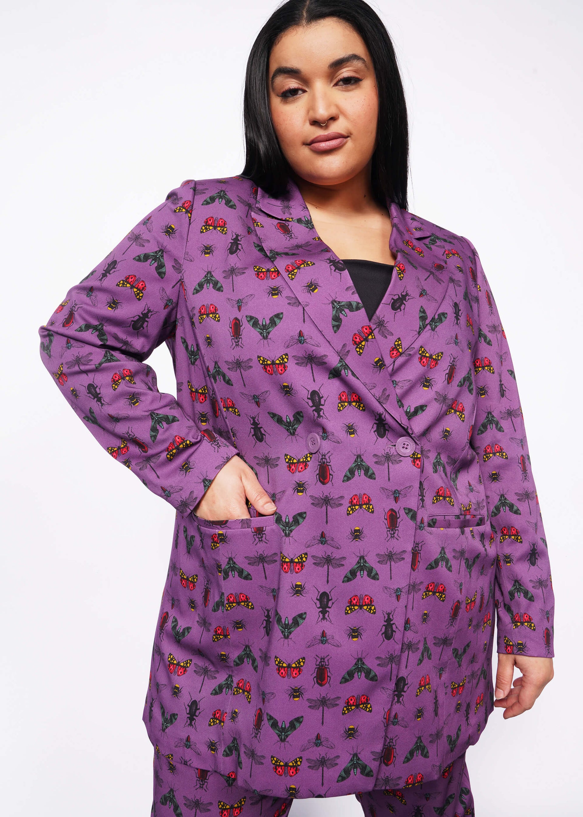 1X=Front view of person with long black hair and nose ring smiling at camera with hand in double breasted blazer pocket. Blazer is buttoned closed and is purple with bug print.