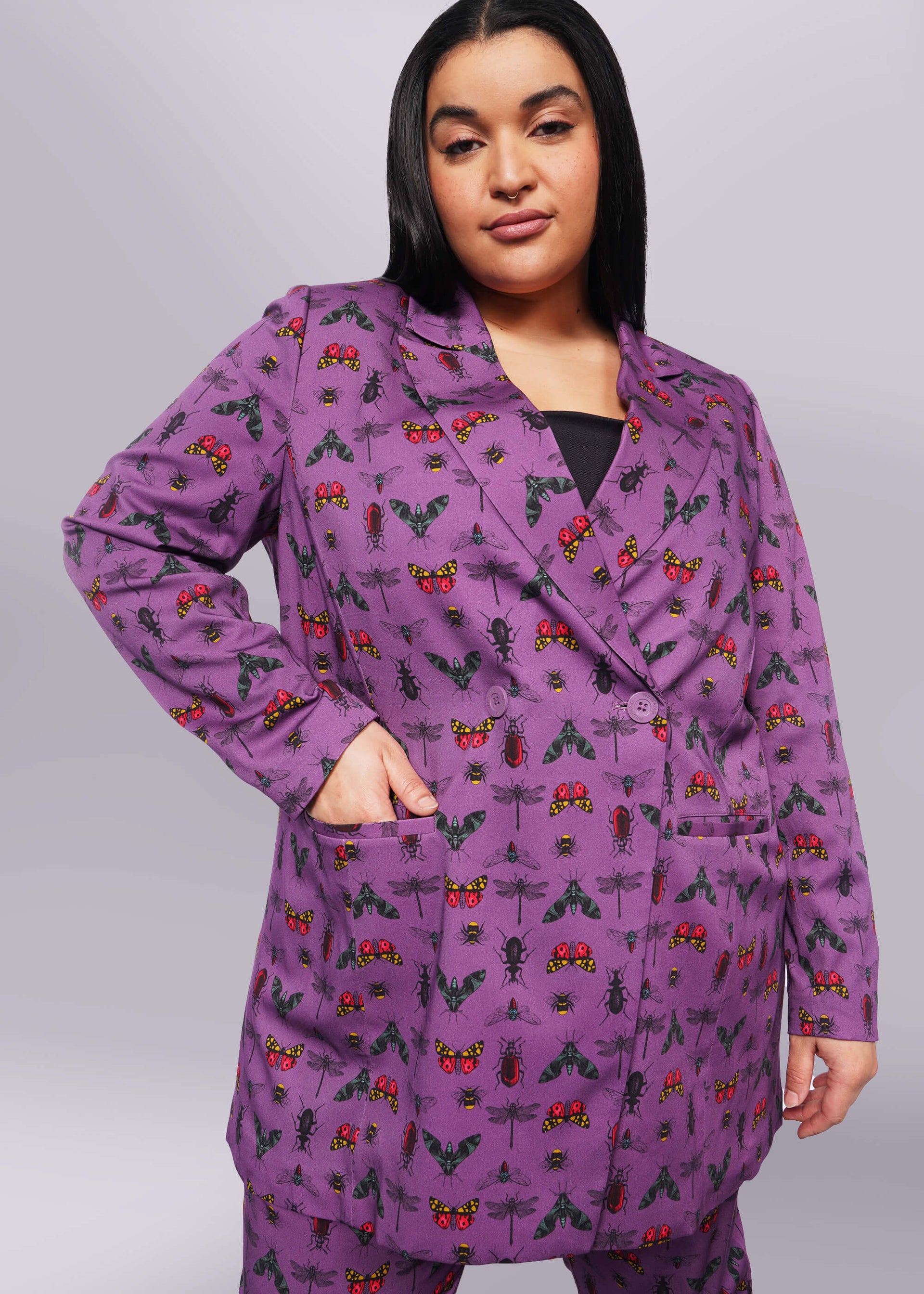 1X=Front view of person with long black hair and nose ring smiling at camera with hand in double breasted blazer pocket. Blazer is buttoned closed and is purple with bug print.