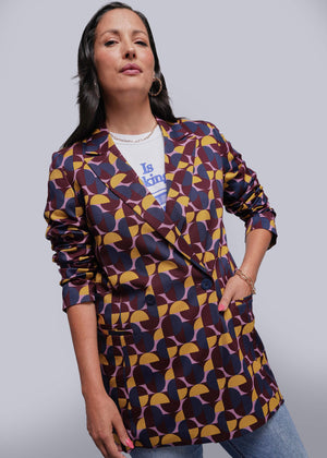 S=Woman wearing a patterned blazer over a white shirt with text, standing against a plain background