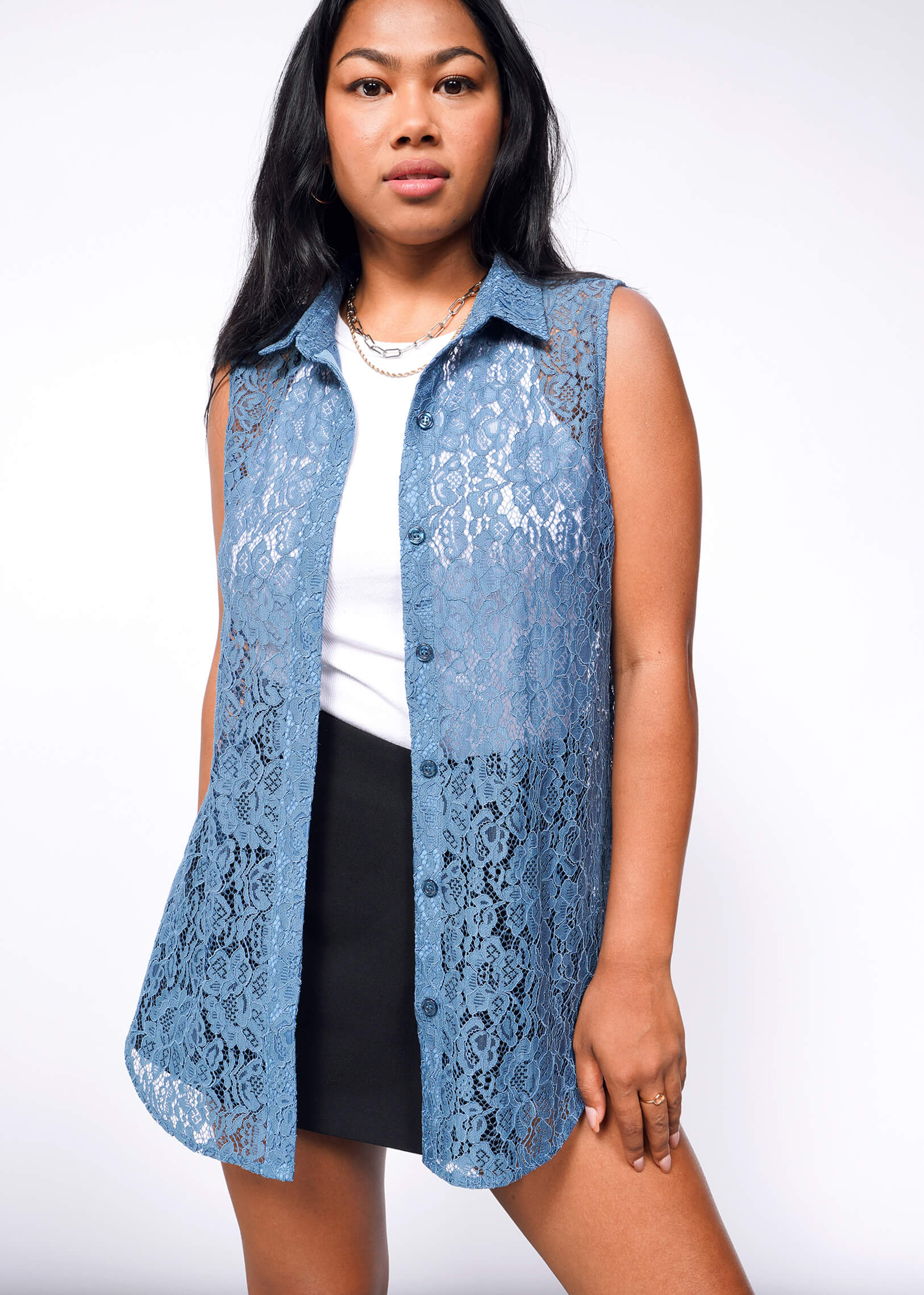 S=A woman poses in The Empower Lace Sleeveless Button Up, a relaxed-fit blue lace top over a white tank and black skirt, against a plain white background with one hand by her side and the other on her hip.