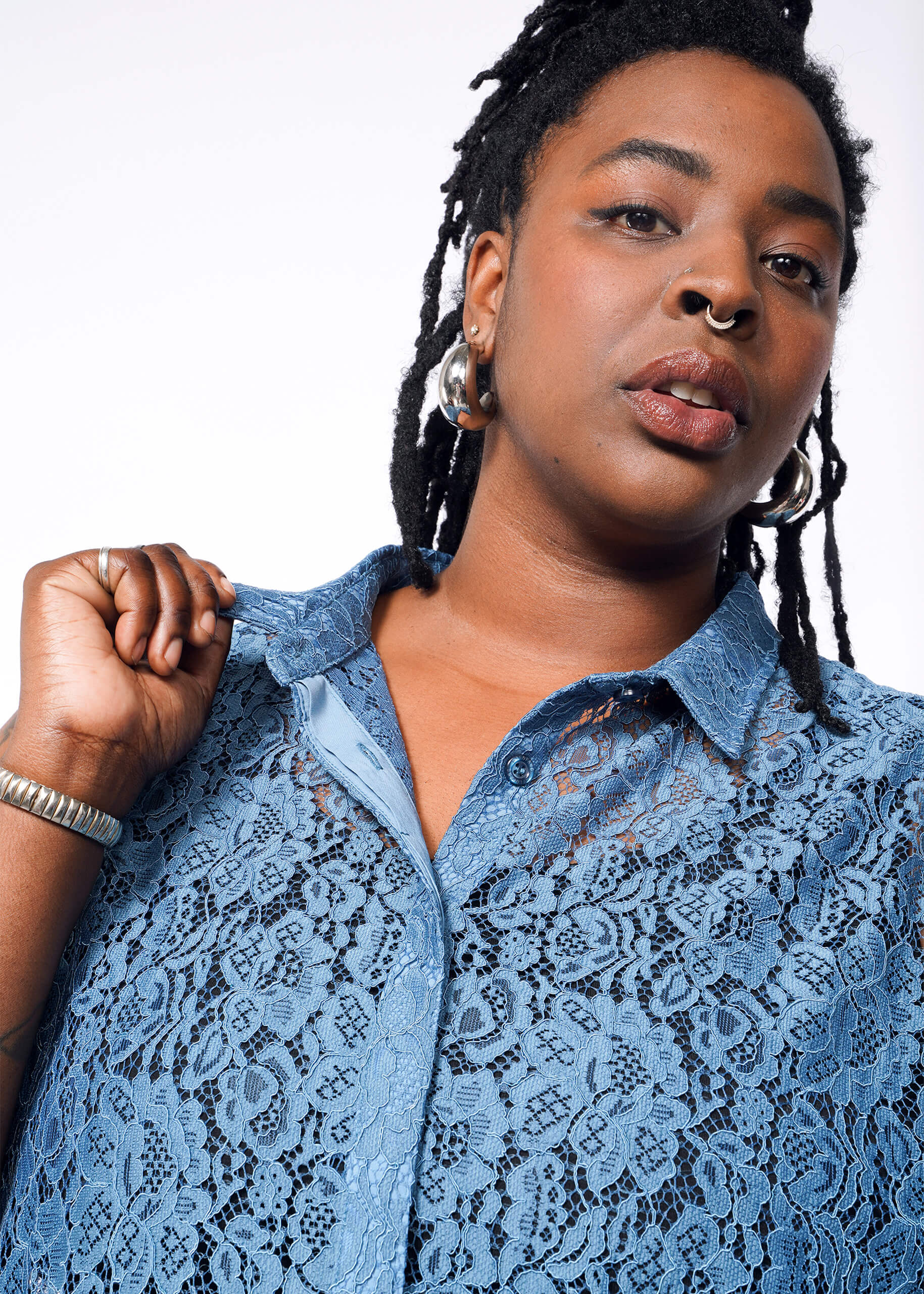 1X=With short dreadlocks and hoop earrings, a person showcases a relaxed vibe in The Empower Lace Sleeveless Button Up, gripping the collar with one hand. A crisp white background accentuates the scene.
