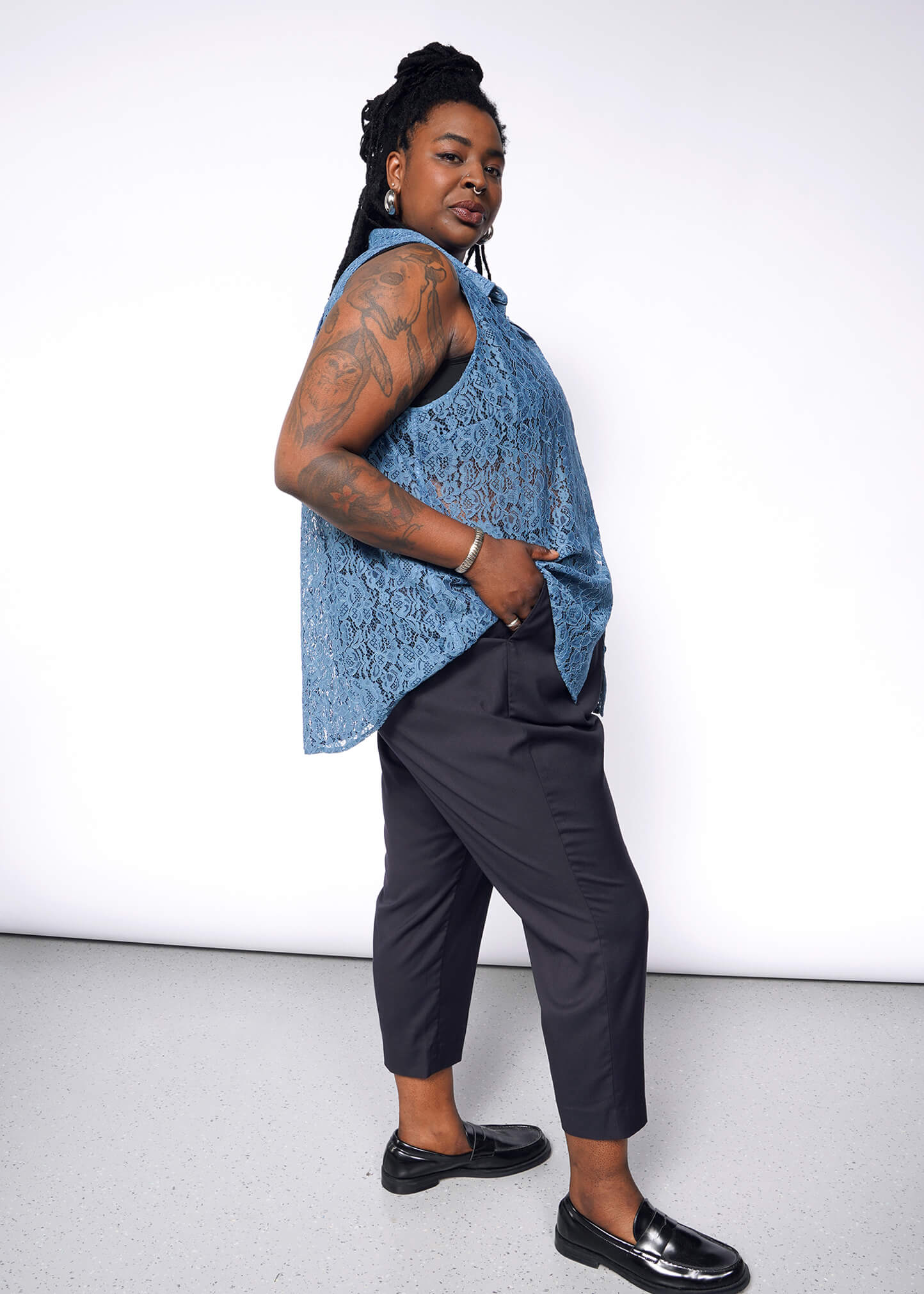 1X=A person with long braided hair stands confidently in The Empower Lace Sleeveless Button Up, featuring a relaxed fit. With tattoos on one arm and hands in pockets, they wear black loafers against a plain light wall.