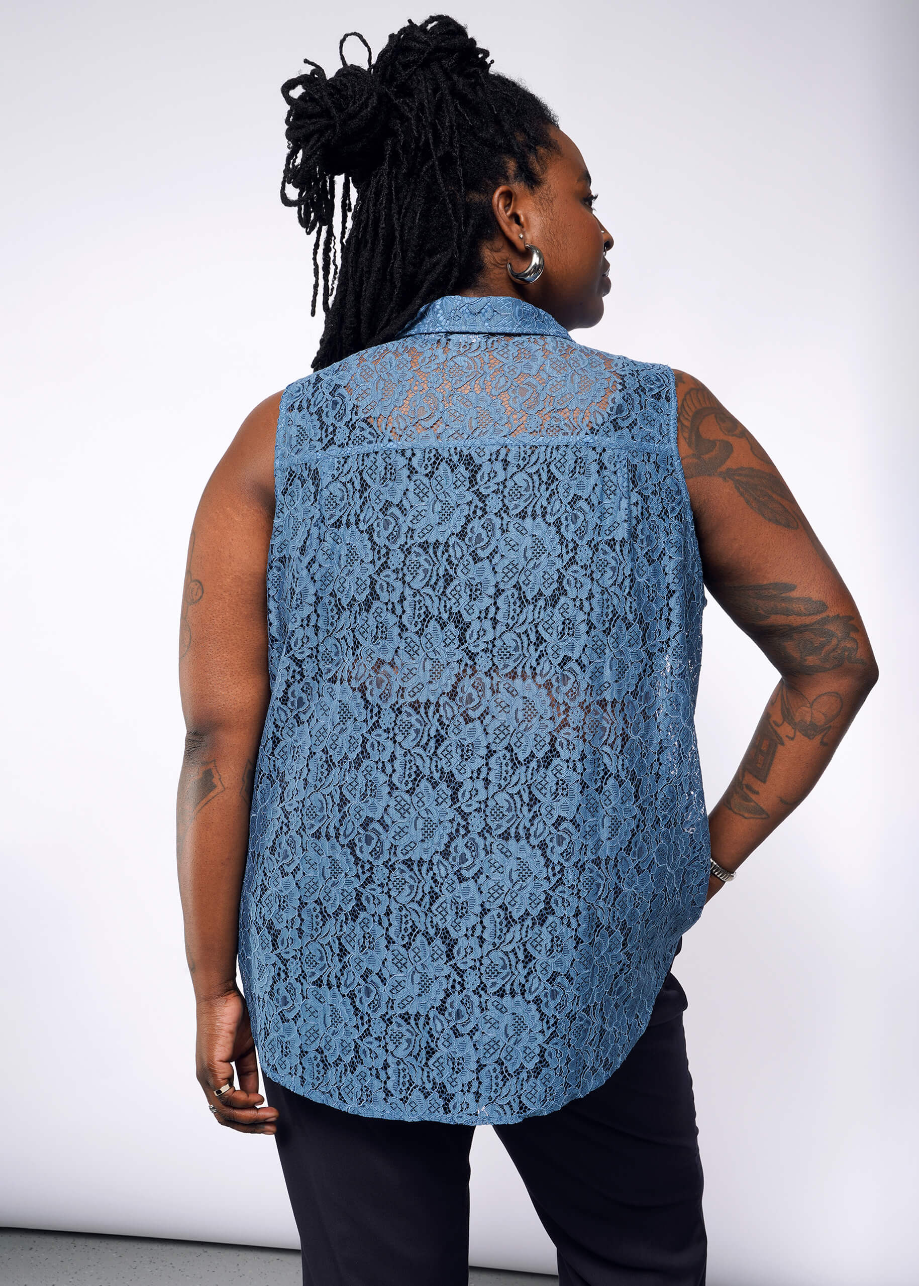 1X=Facing away, a person with long braided hair wears The Empower Lace Sleeveless Button Up and relaxed-fit dark pants. Tattoos adorn their arms against a plain, light-colored background.