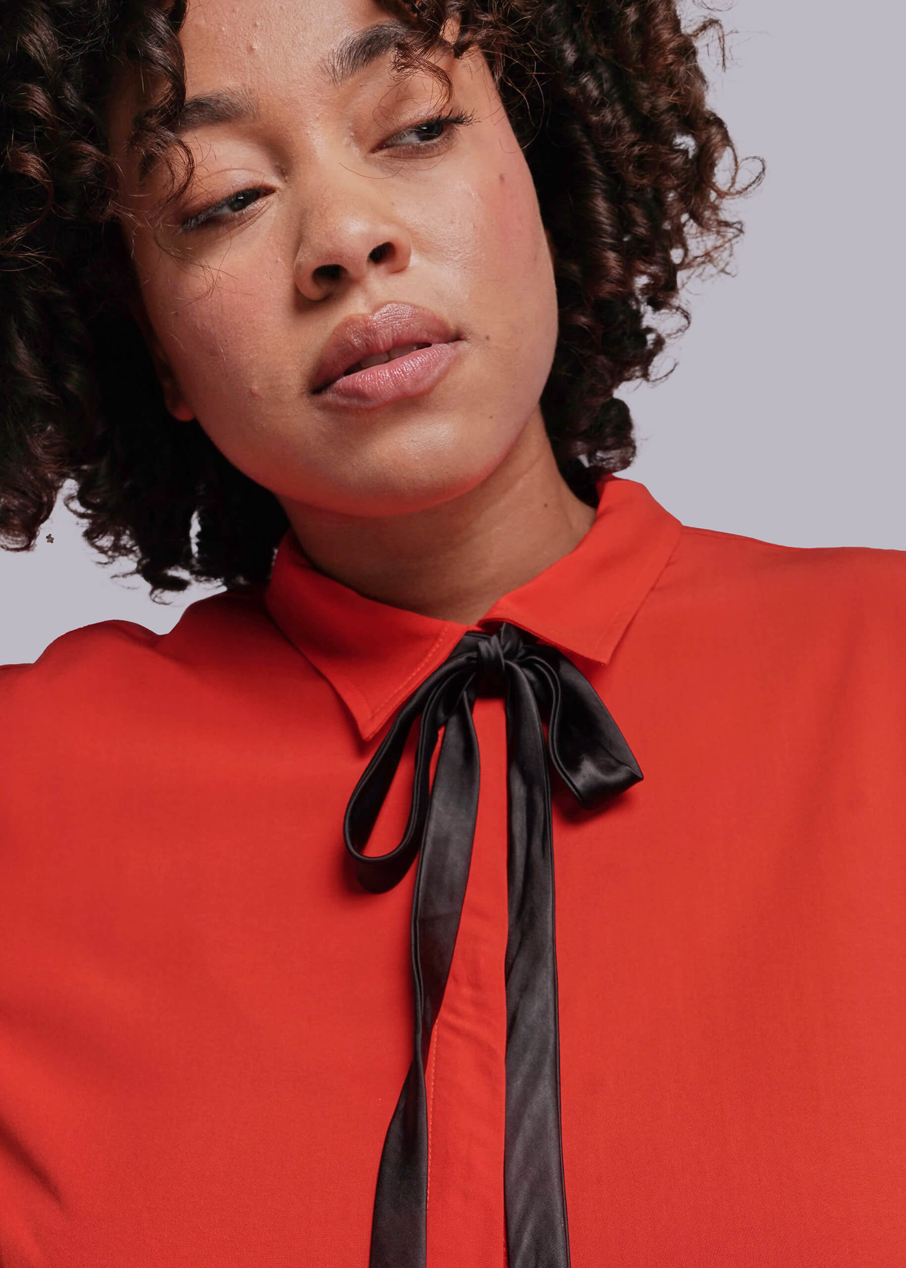 1X=Person wearing a red blouse with a black ribbon tie against a plain background