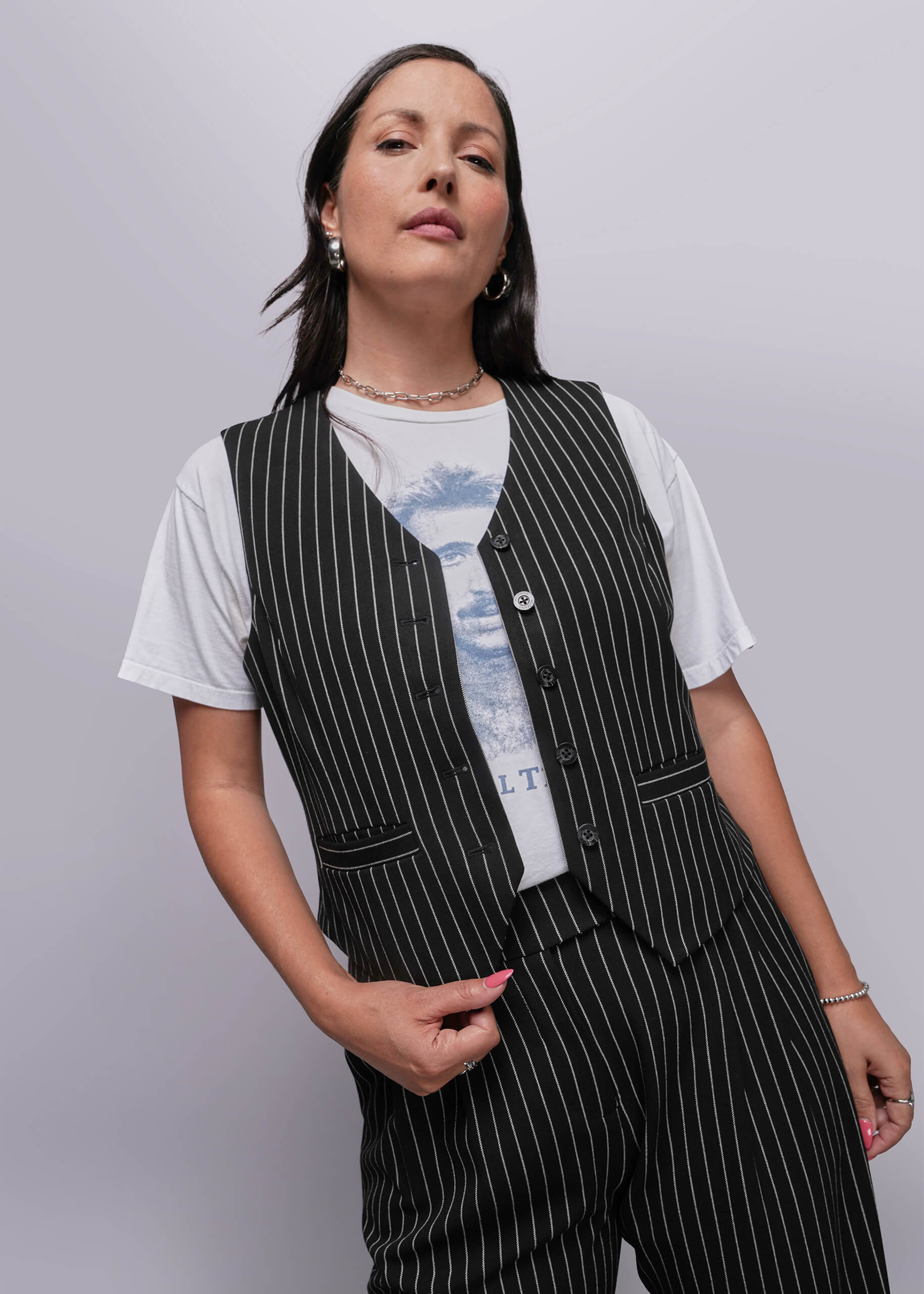 1X=Woman wearing a black pinstripe vest over a white t-shirt with a graphic design on a plain background