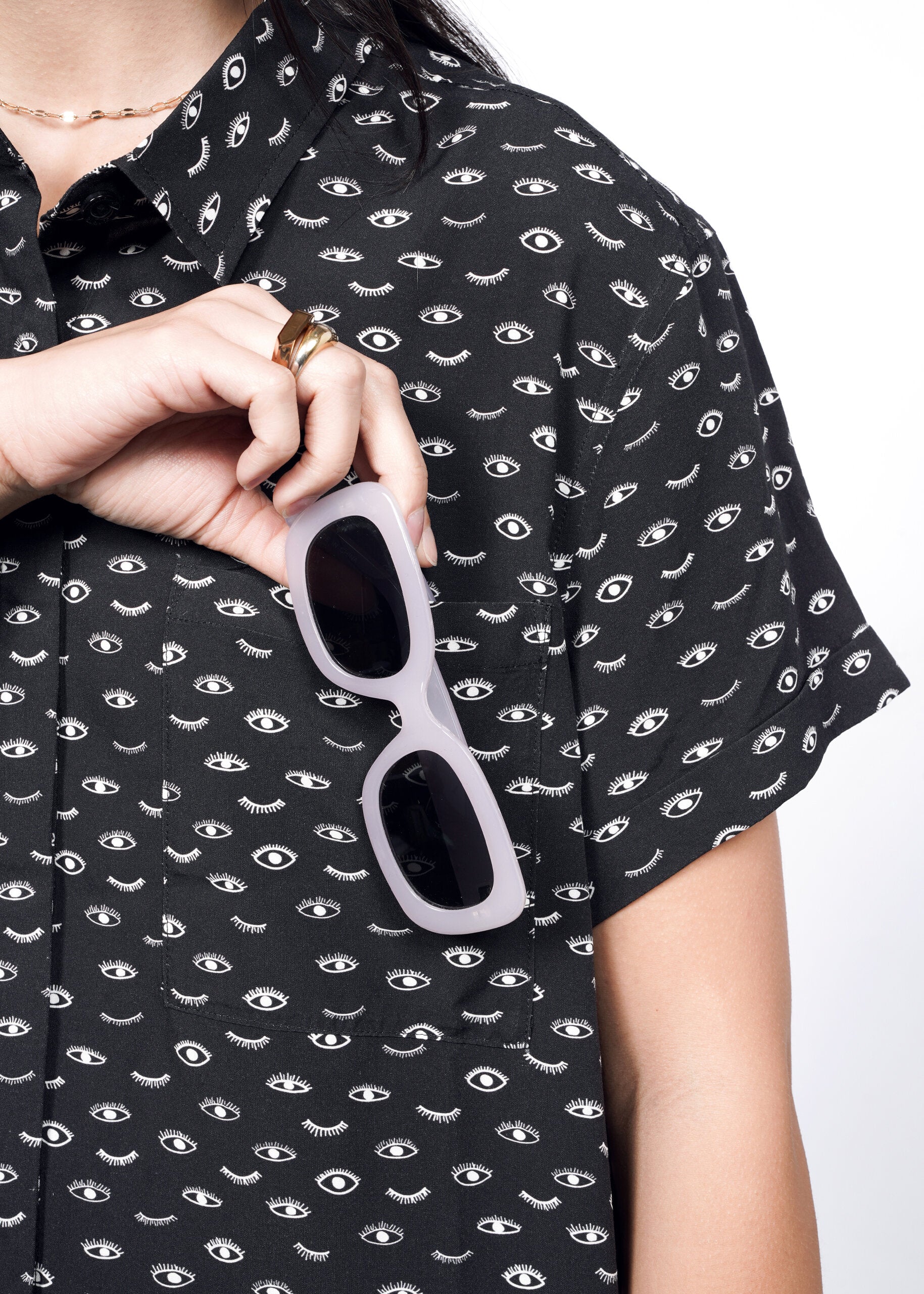 S=A person in The Empower Shirt Dress, featuring a black design with a white eye pattern, holds white-framed sunglasses near their chest. Only part of the torso is seen against a plain white background.
