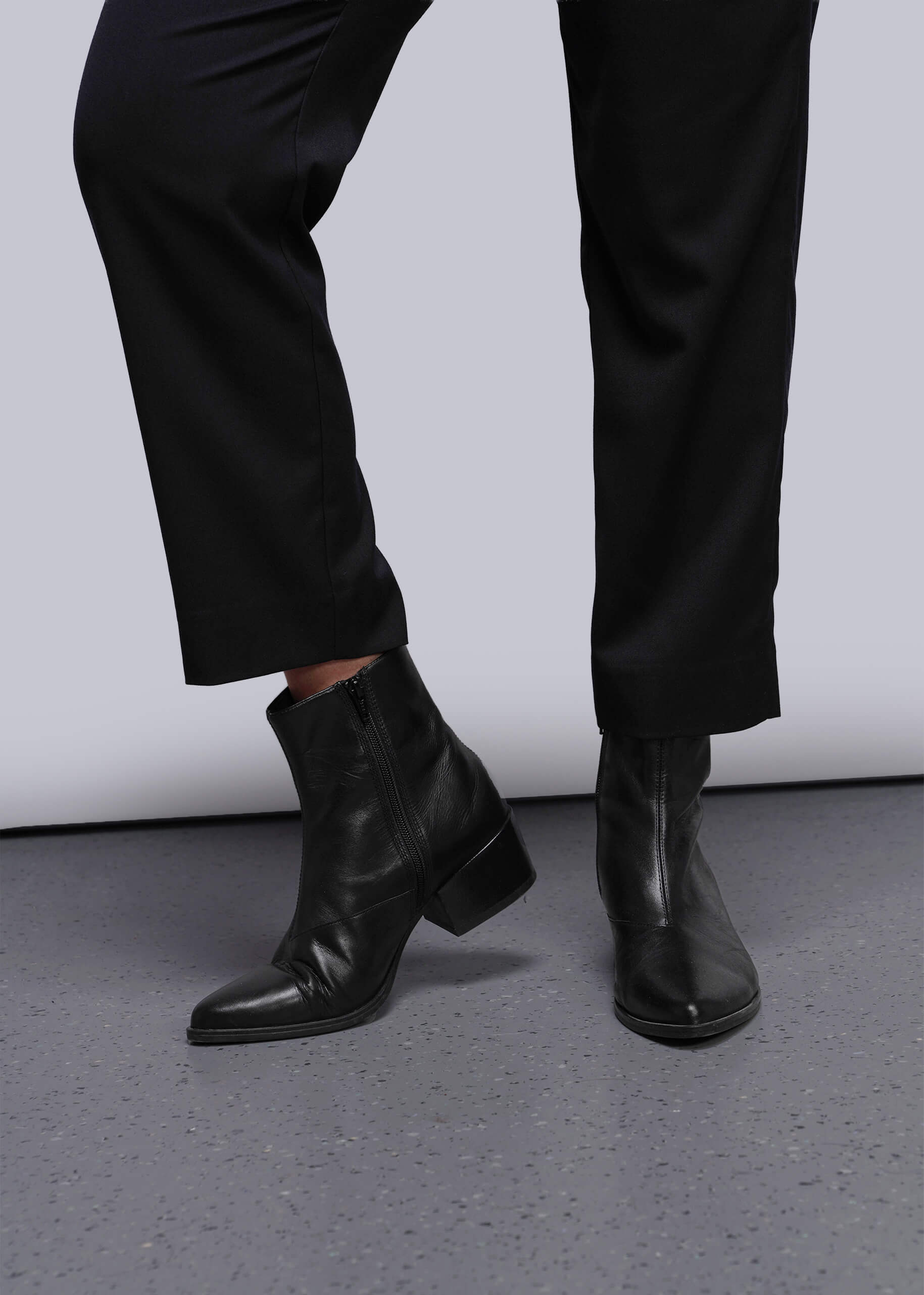 S=Black leather ankle boots worn with black pants on a gray floor.