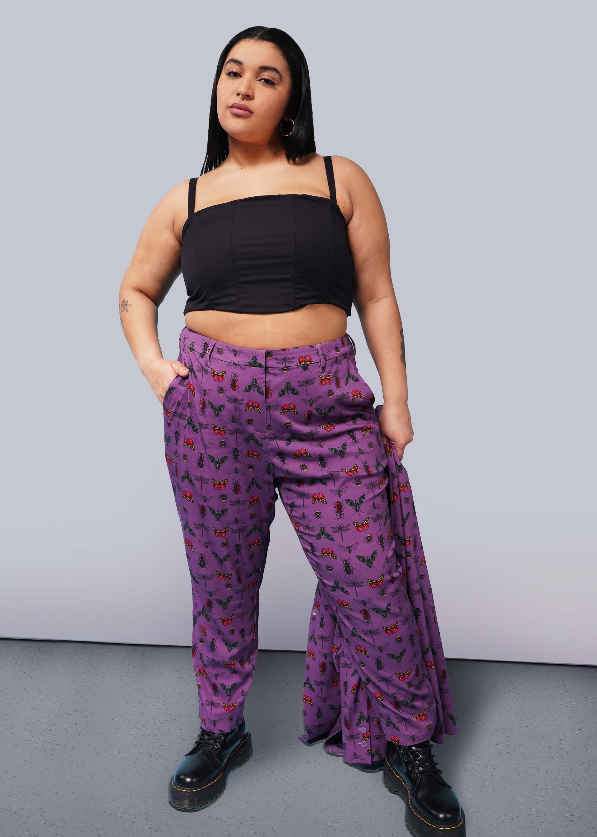 1X= Person with long black hair looks at camera with hand in pocket wearing black corset, purple bug printed slim crop pants, and Doc Marten boots.